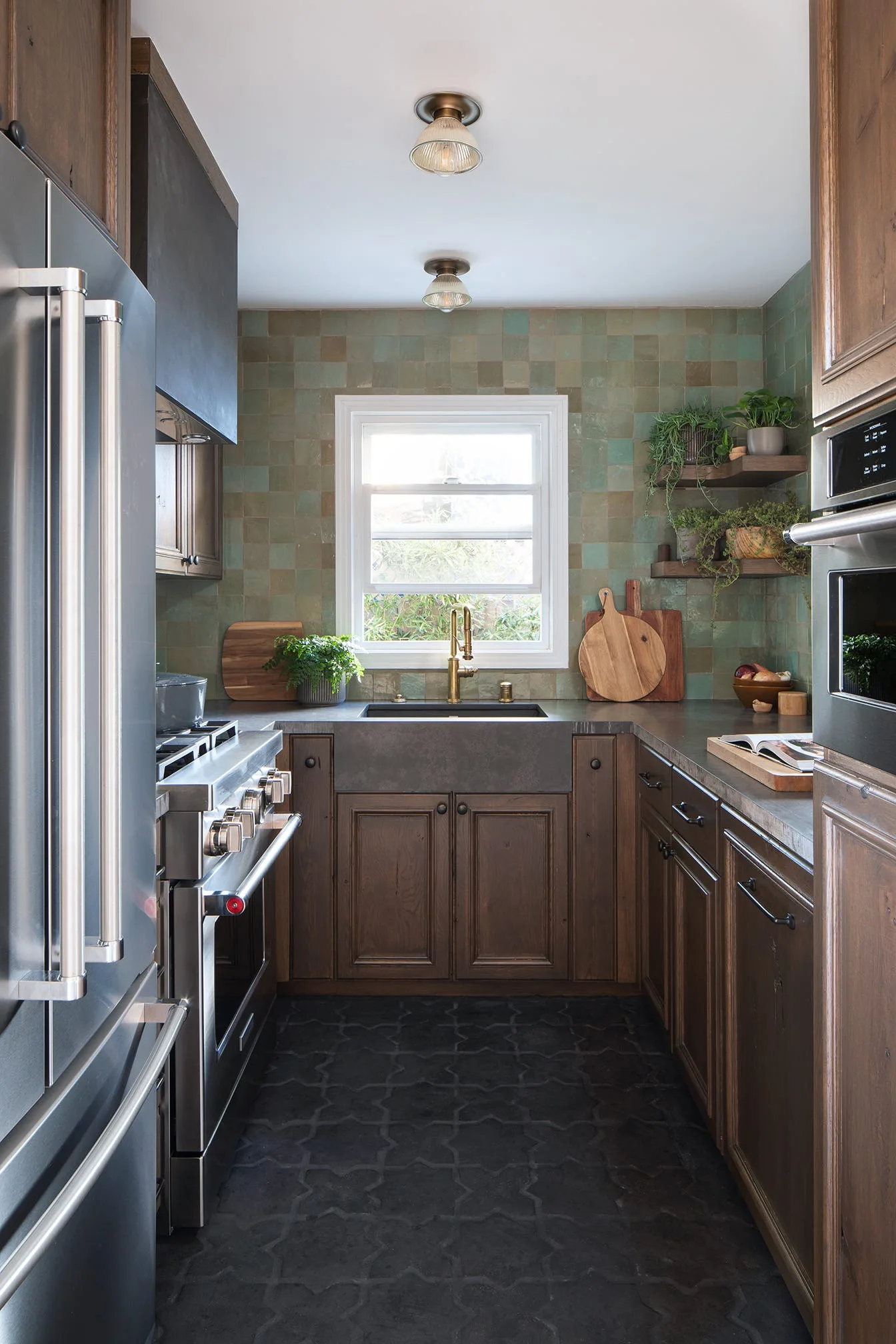 A narrow galley kitchen featuring dark wood cabinets, a green tiled backsplash, a deep farmhouse sink, and stainless steel appliances.
