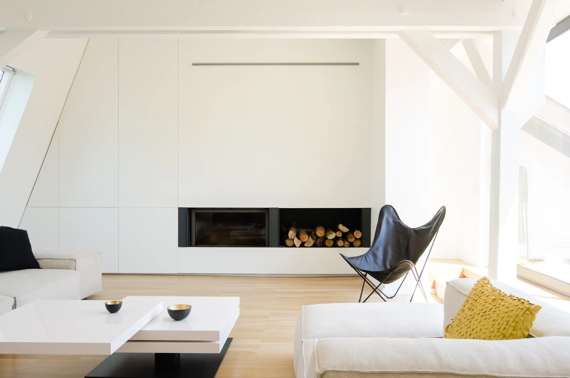 A bright attic living room featuring a modern black fireplace built into a flat white wall, a white sofa, and a black leather butterfly chair.