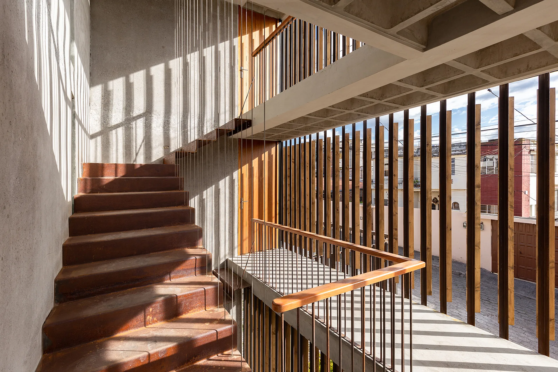 A modern industrial stairwell features a rusted metal staircase, concrete walls, and a vertical wood slat screen filtering natural light.