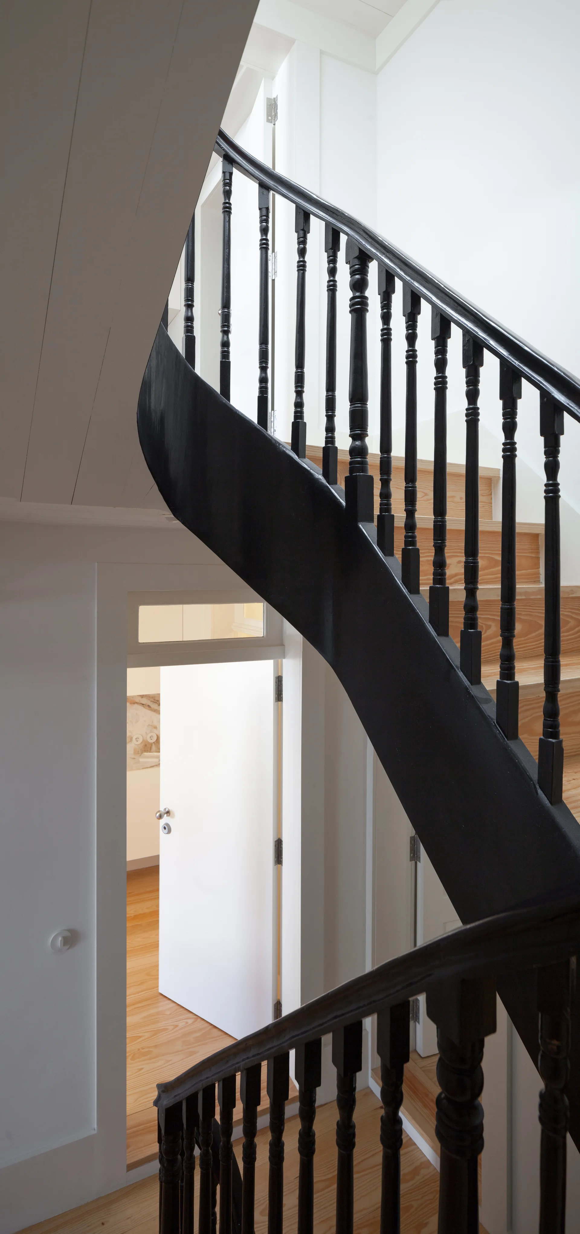 A curved wooden staircase features black painted railings and balusters against clean white walls with a view looking down into a bright hallway.