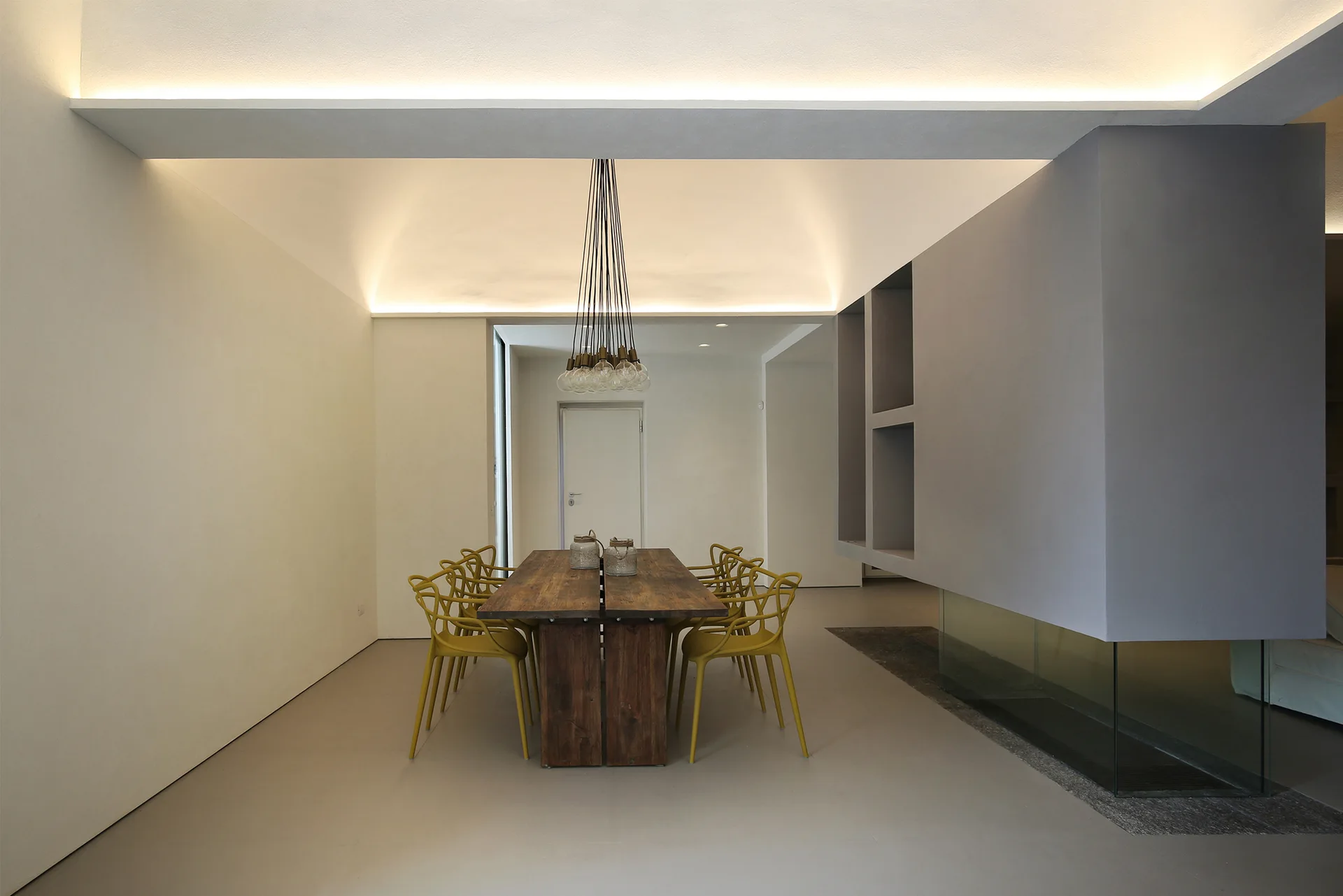 A minimalist dining room featuring a rustic wood dining table and yellow chairs illuminated by warm ceiling cove lighting.
