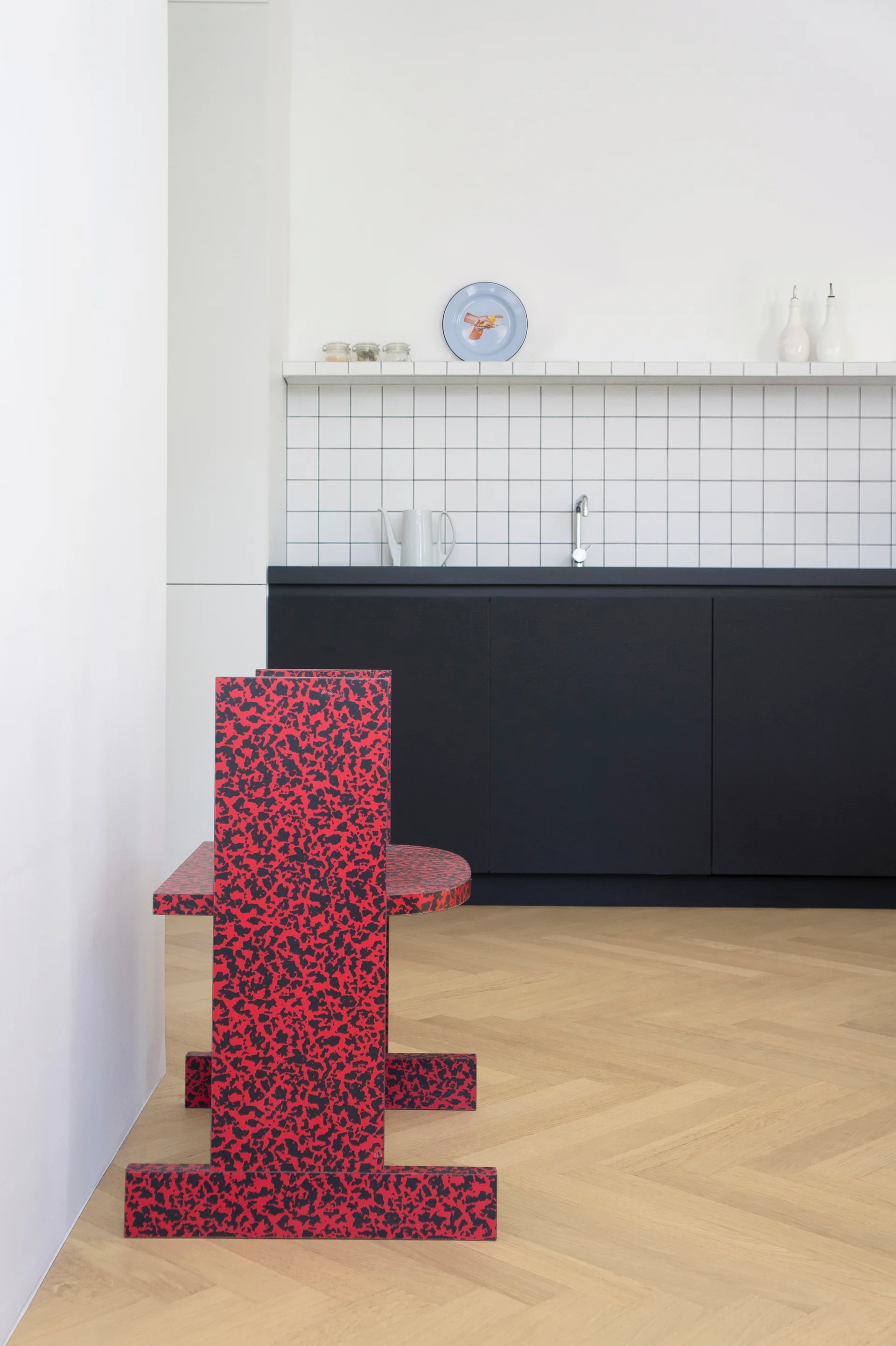 A red and black speckled chair sits on a herringbone wood floor in a modern kitchen with matte black lower cabinets and a white tiled backsplash.
