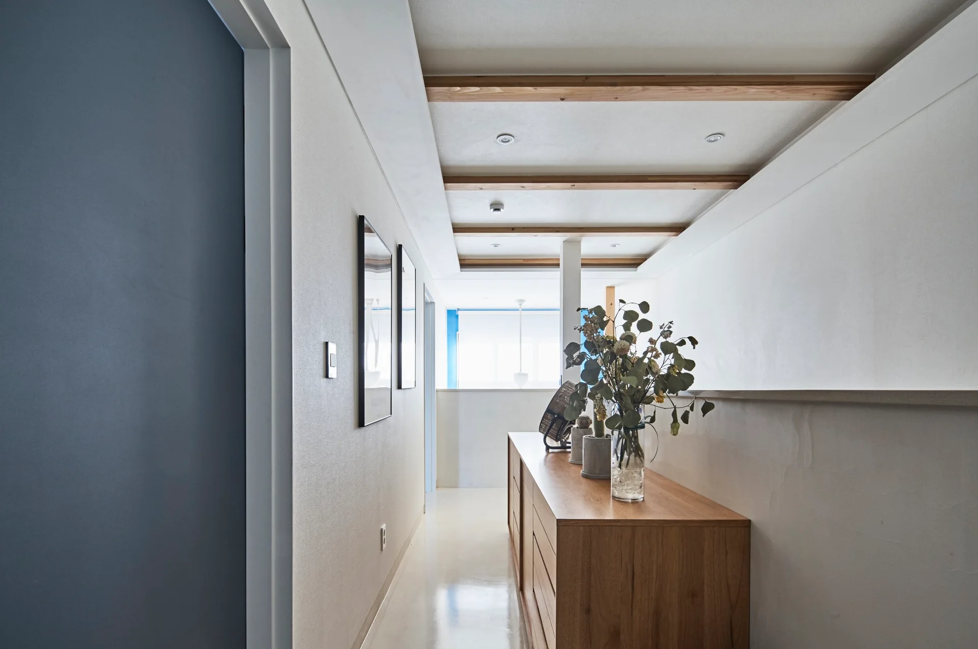 A modern hallway features a wooden console table with a glass vase of branches underneath a ceiling with exposed wood beams.