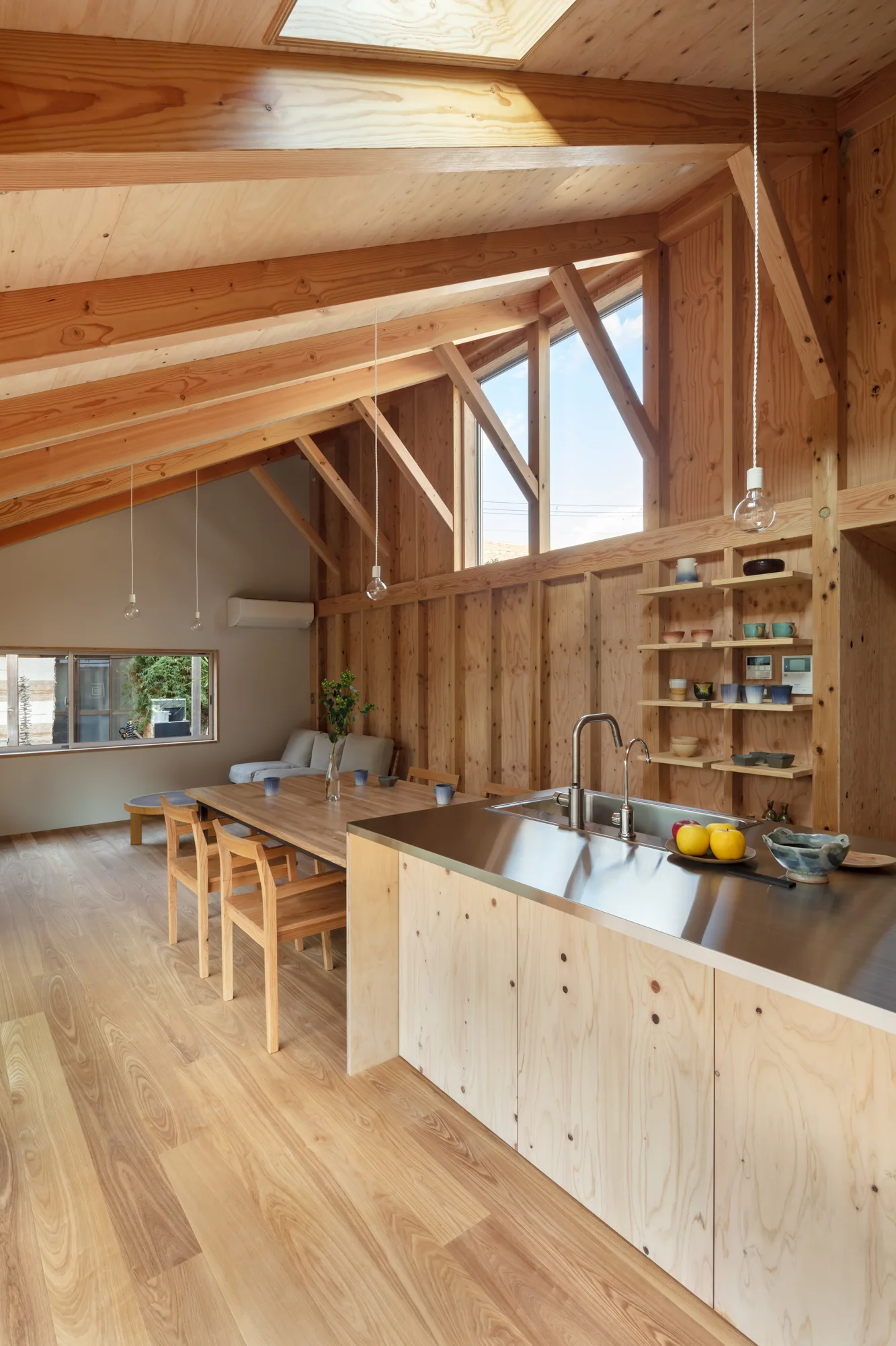 A minimalist kitchen and dining space with a plywood island, stainless steel countertop, wooden dining set, and vaulted ceiling with exposed beams.