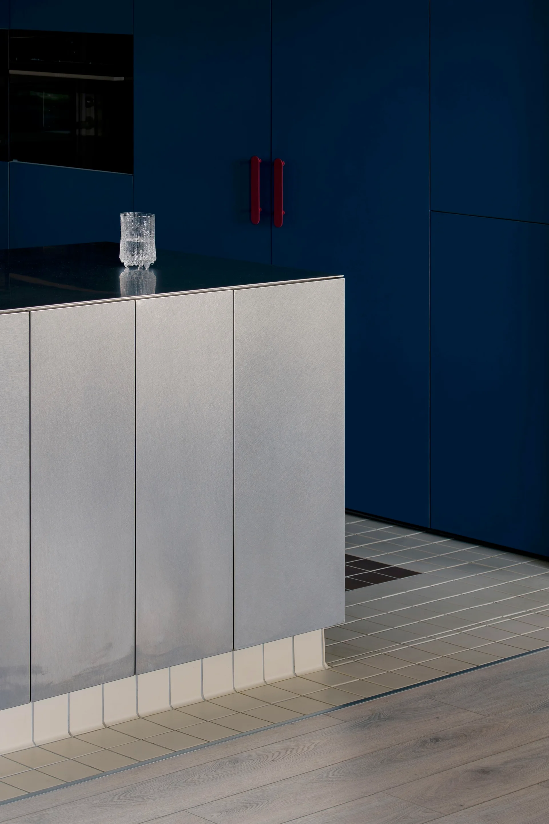 A modern kitchen island with satin metal paneling stands before deep blue cabinets and a tiled floor transition.