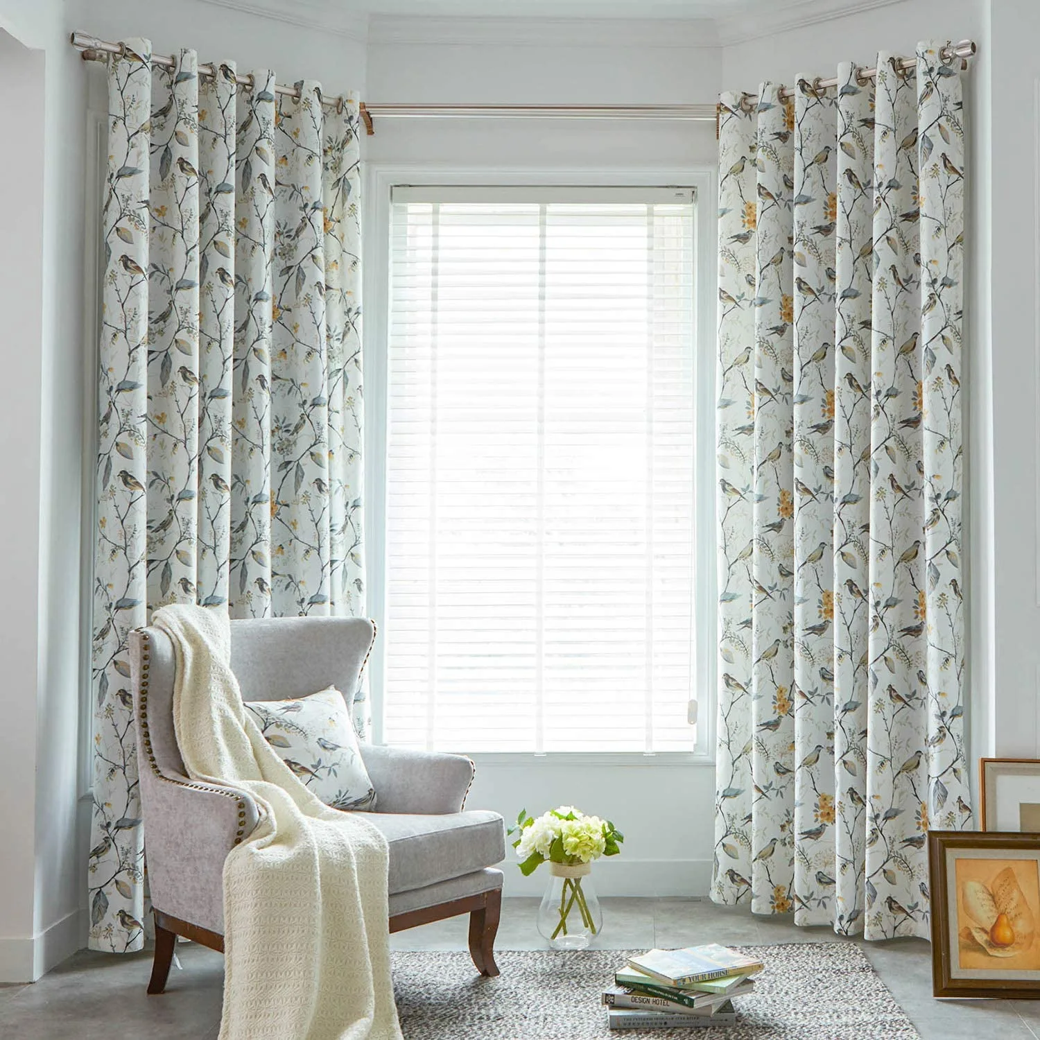 A light gray wingback armchair with a cream throw sits by a bay window dressed in bird-patterned curtains and white venetian blinds.