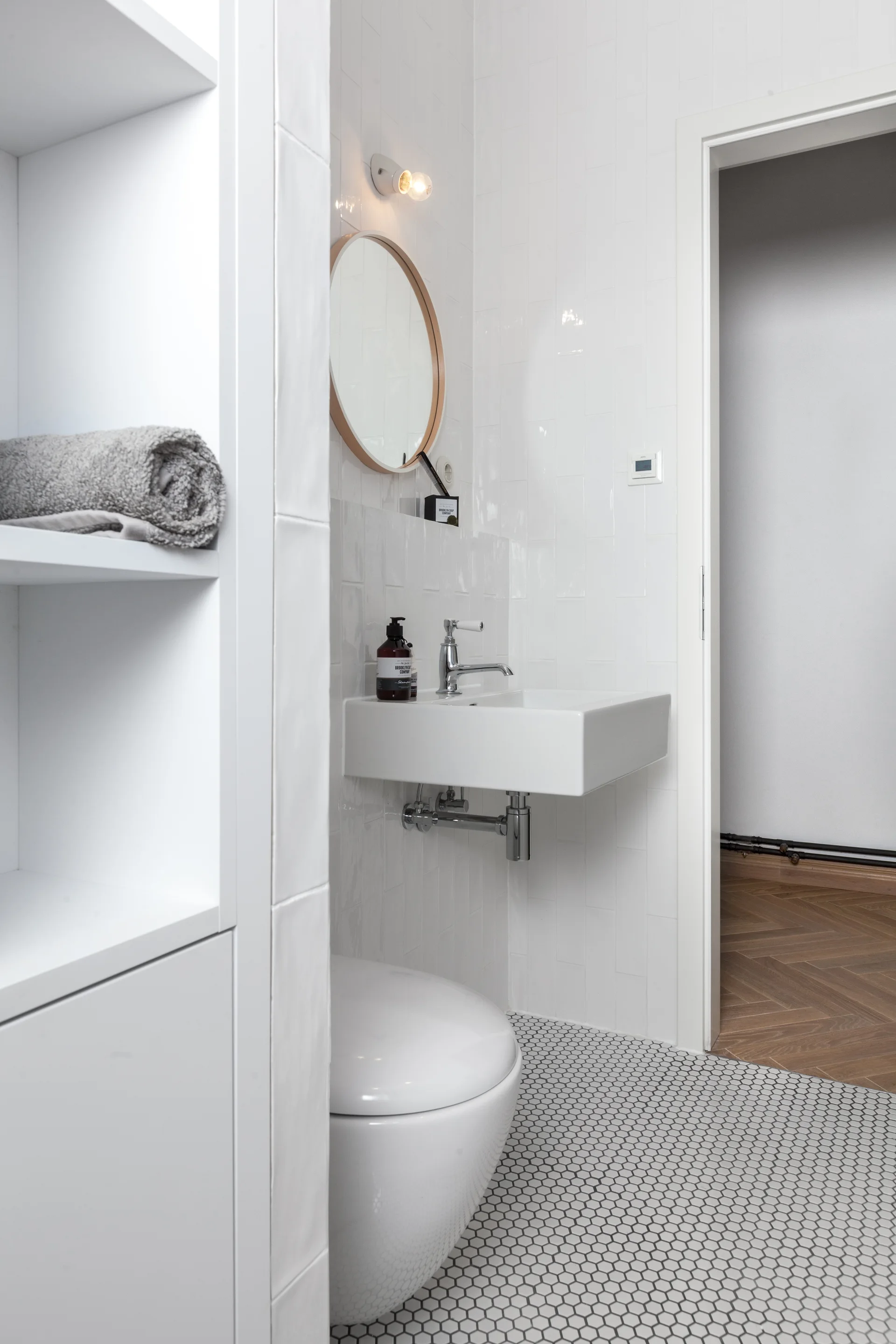 A small white tiled powder room features a wall-mounted sink, a round wood-framed mirror, a wall-mounted toilet, and small hexagonal mosaic floor tiles.