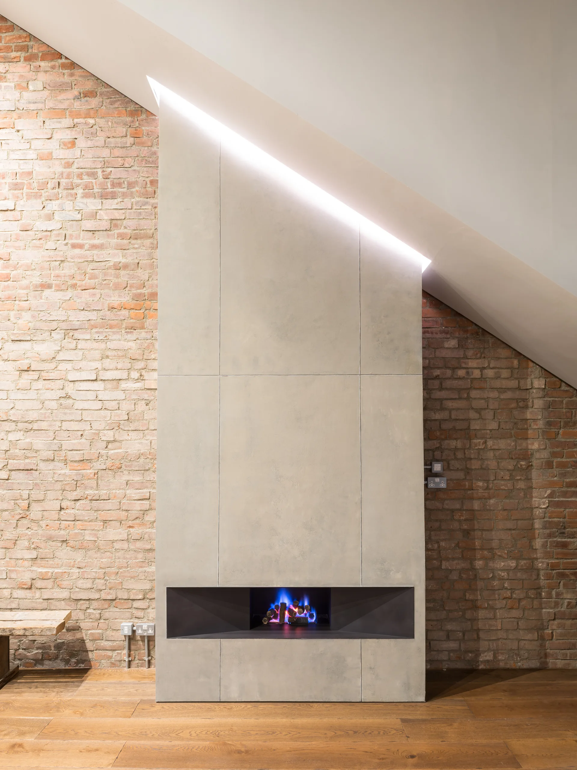 A grey concrete fireplace structure built against a rustic exposed brick wall under a sloped white ceiling with recessed linear accent lighting.