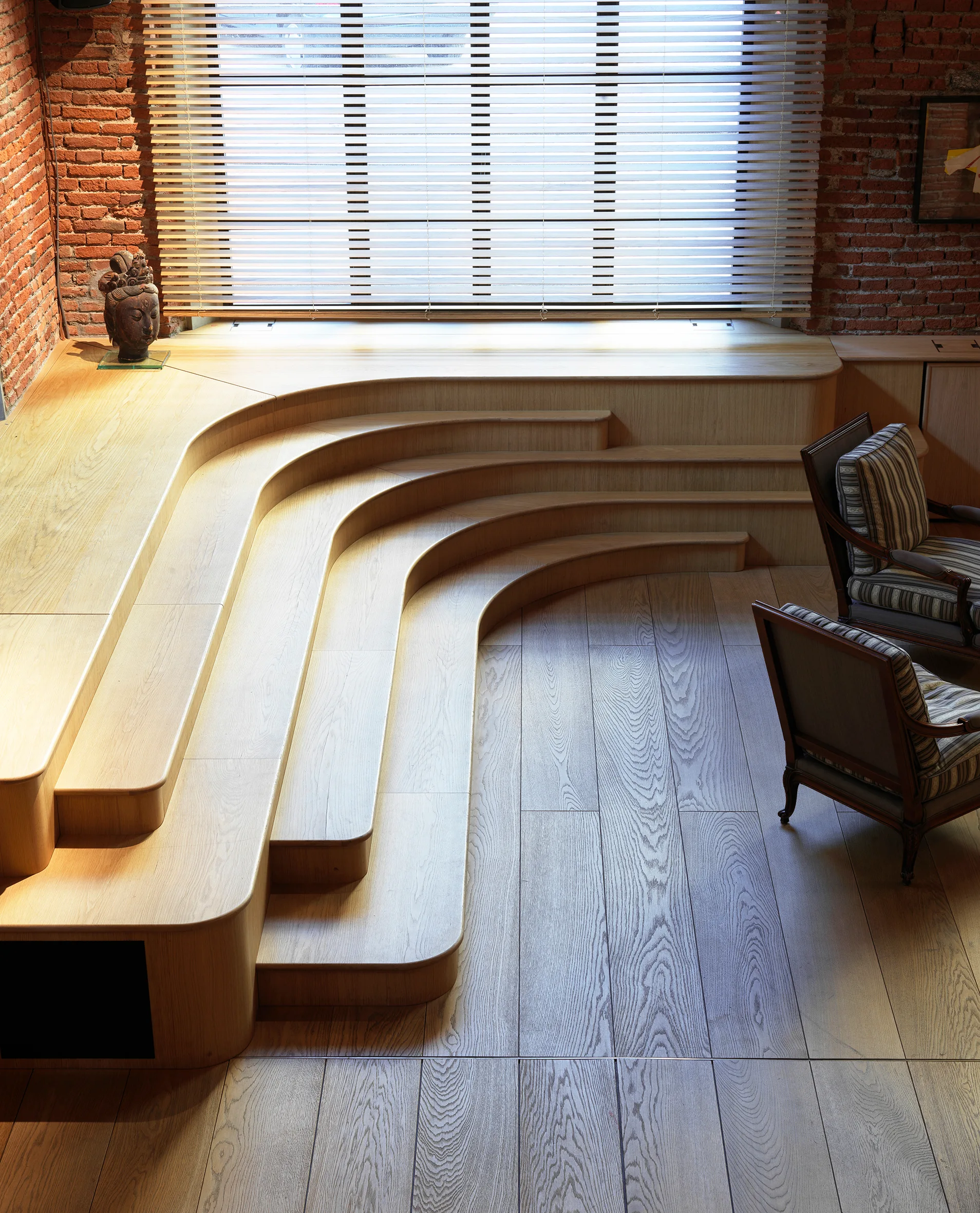 A light oak custom stepped platform curves along an exposed brick wall next to two wooden armchairs in a loft living space.