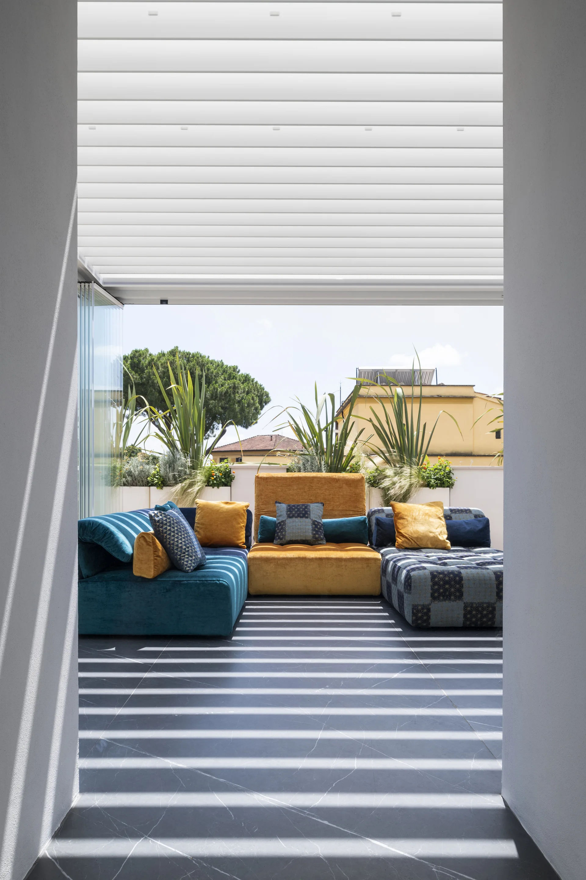 A view through a doorway shows a contemporary terrace with a colorful teal and yellow modular sofa beneath a slatted white pergola.