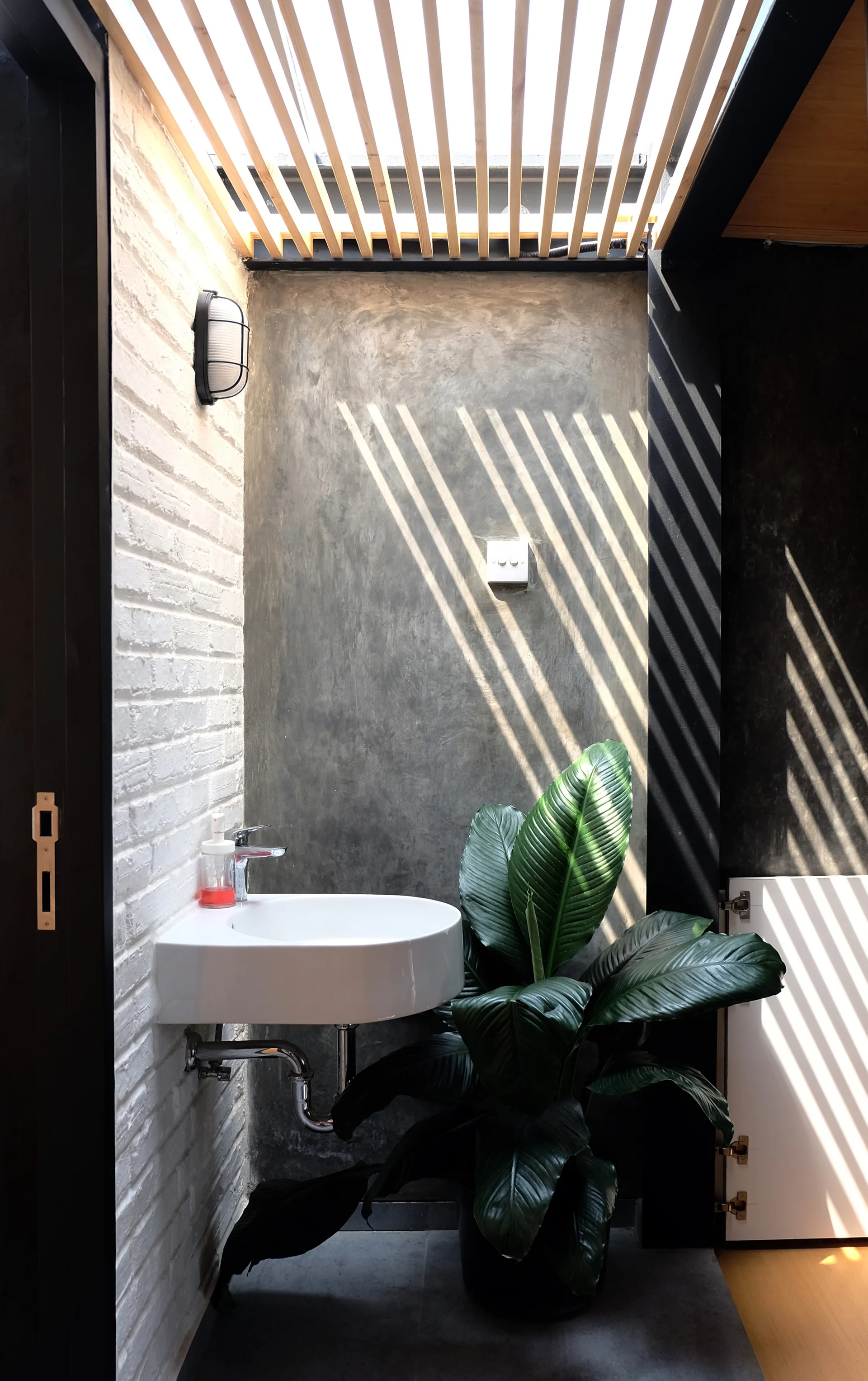 A wall-mounted white ceramic sink is set against a white brick wall and a grey concrete wall under a slatted ceiling that filters natural light.