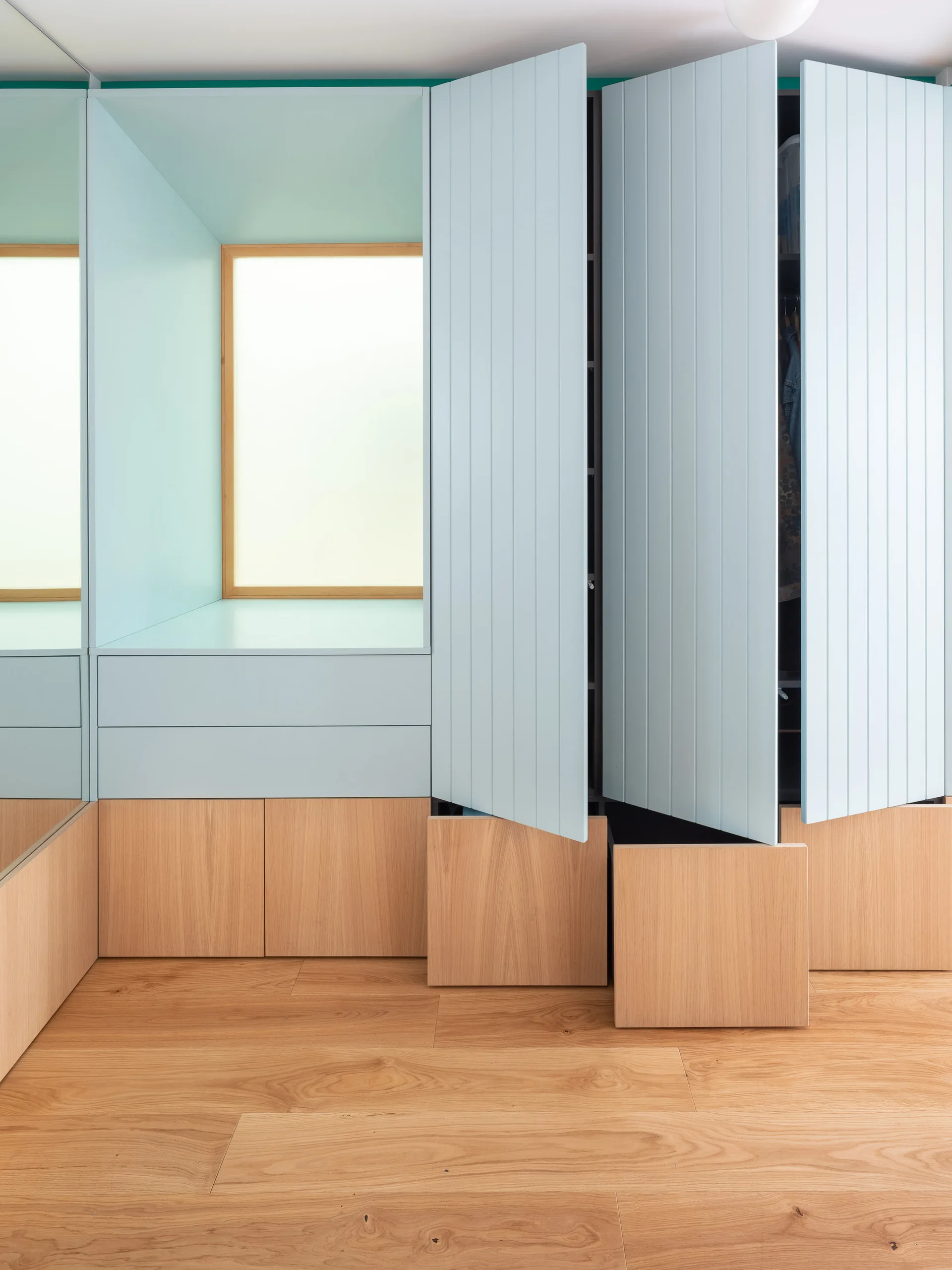 A custom built-in wardrobe features pale blue vertical-planked doors above natural wood drawers next to a mirrored wall and window niche.