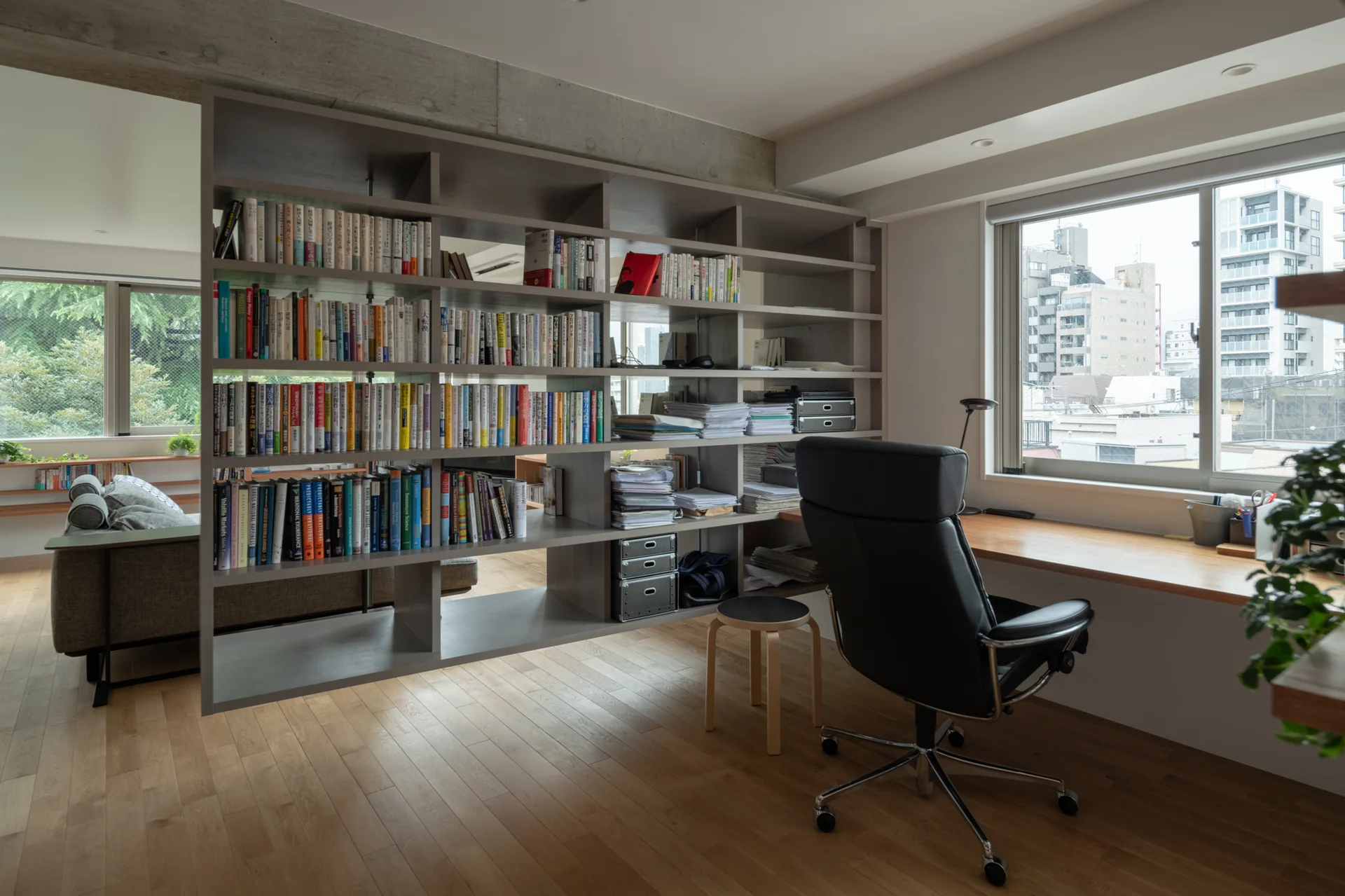 A home office features a wood desk under a window next to a large open bookshelf partition that separates the workspace from a living room.