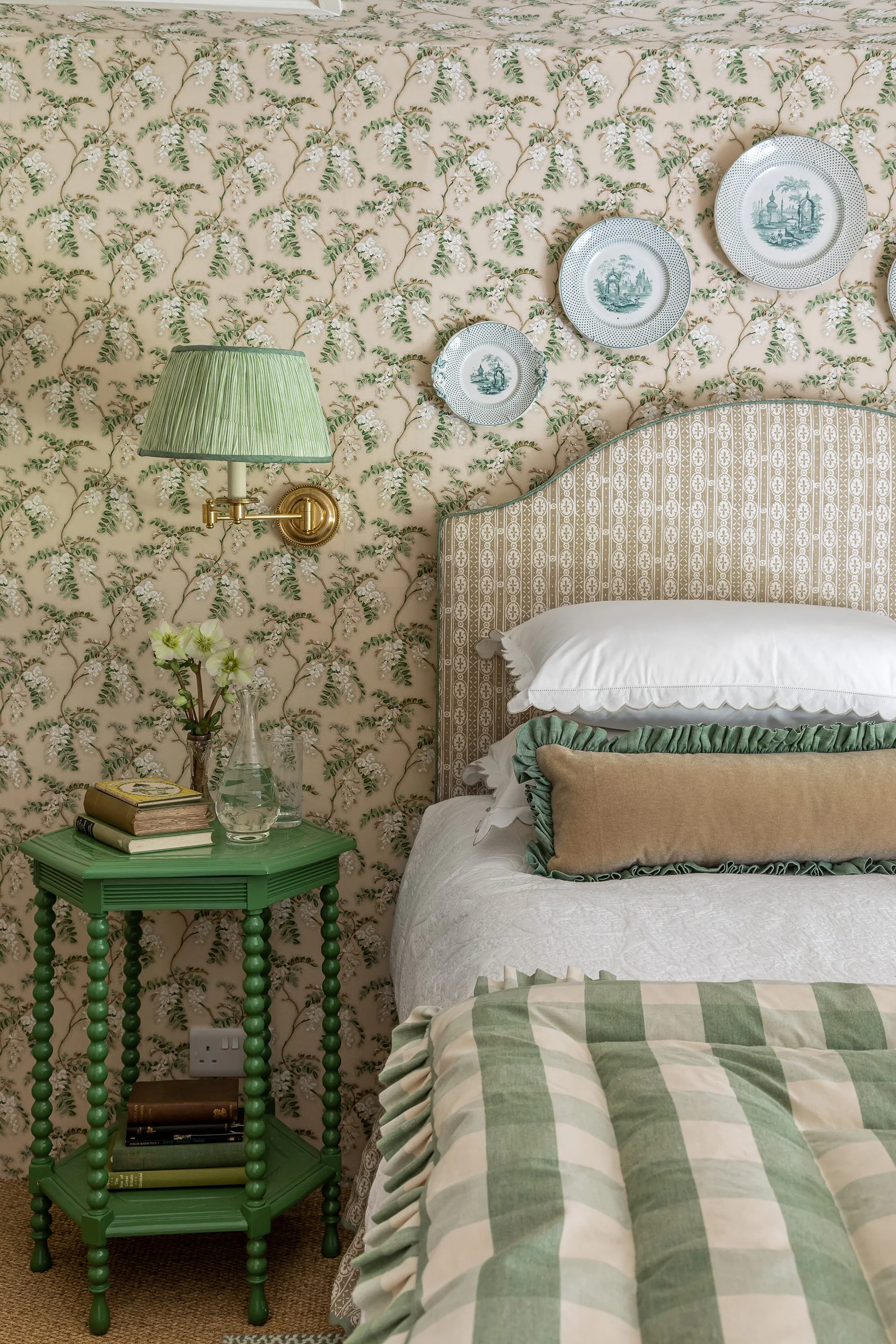 A traditional bedroom features a green bobbin-leg bedside table next to an arched upholstered headboard against trailing floral wallpaper.
