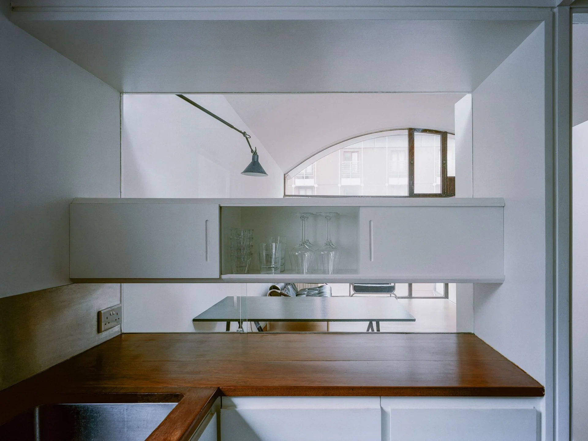 A minimalist white kitchen pass-through with a wooden countertop looks into a dining room with an arched window.