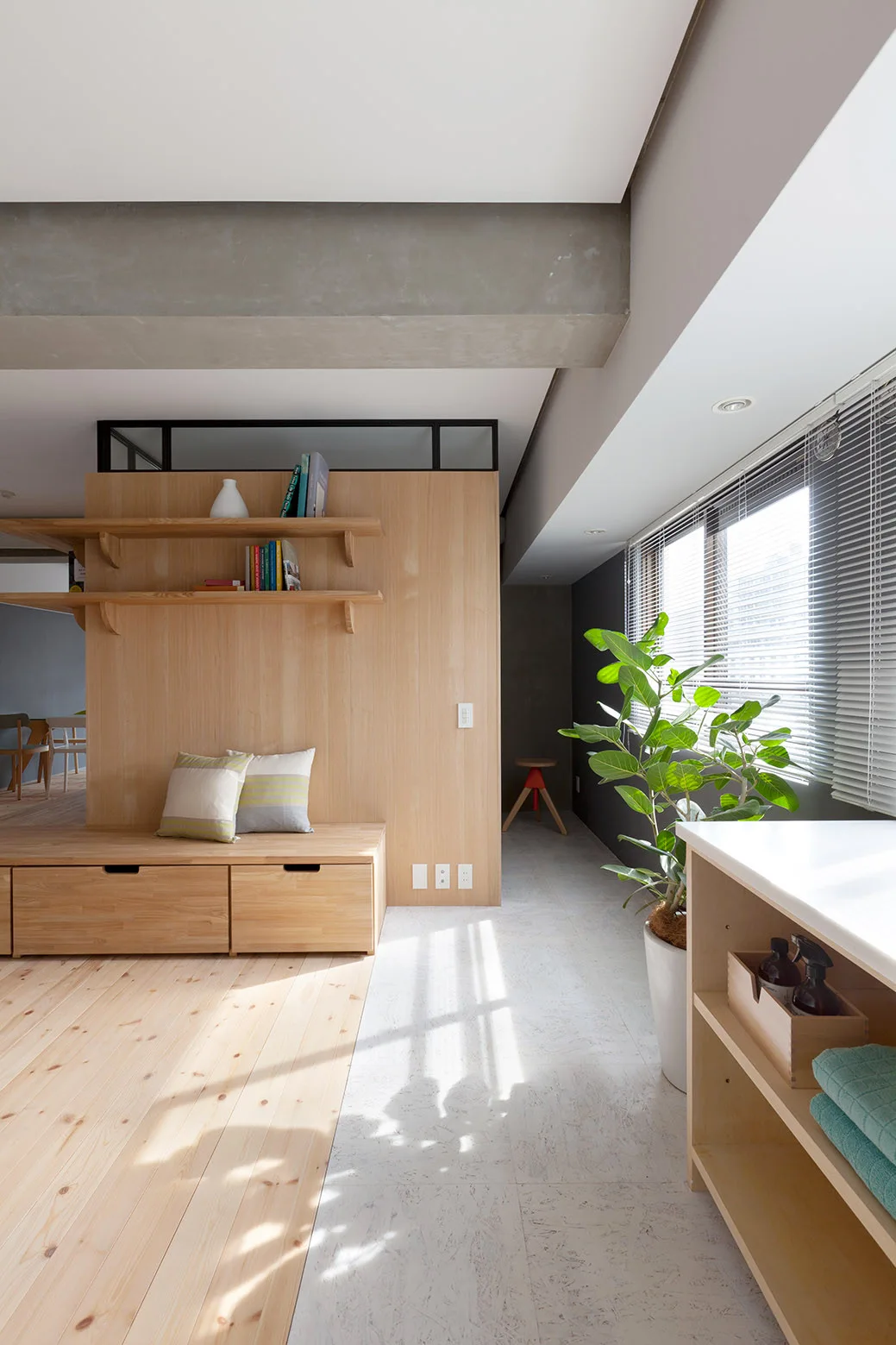 A minimalist room featuring a light wood partition wall with integrated shelving and a storage bench beneath a raw concrete ceiling beam.