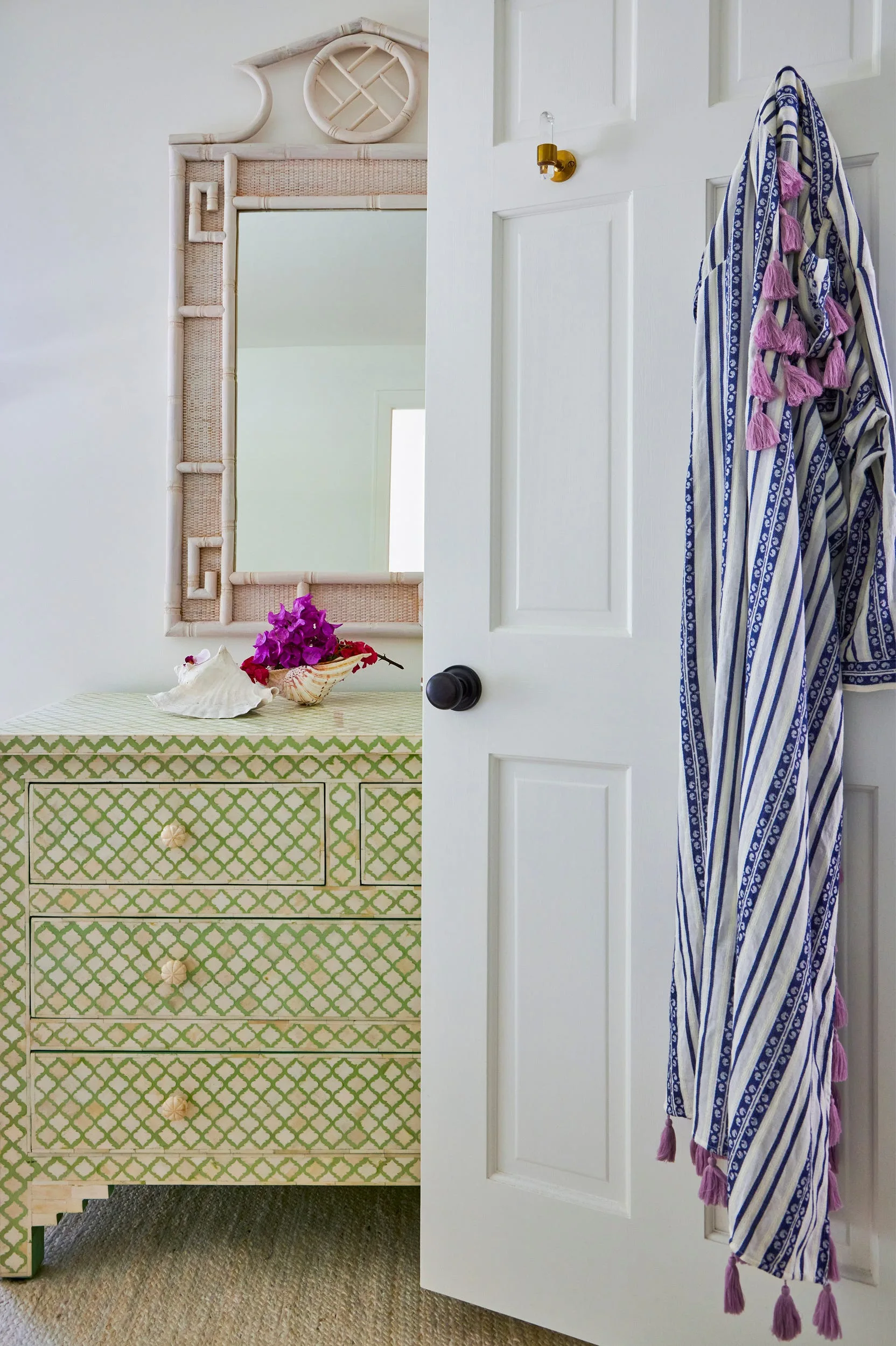 A green and white lattice-patterned dresser sits beneath a bamboo mirror next to an open door hanging a striped robe with purple tassels.
