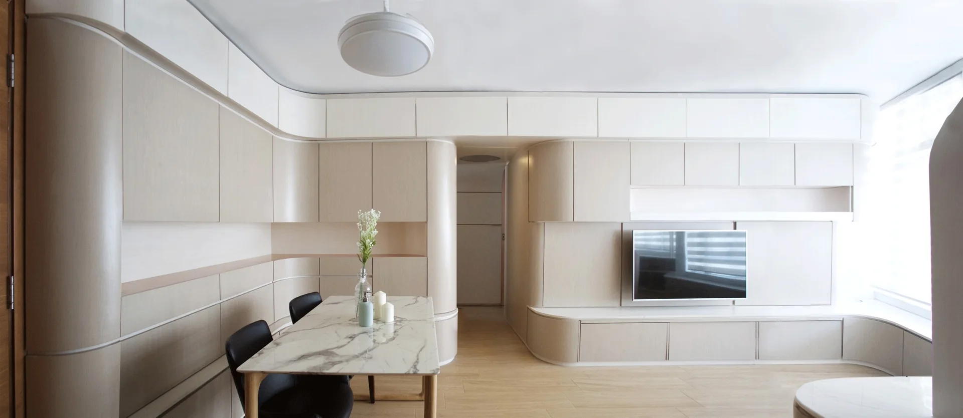 A modern dining and living space features seamless curved beige cabinetry, an integrated flat-screen television, and a marble-topped dining table.