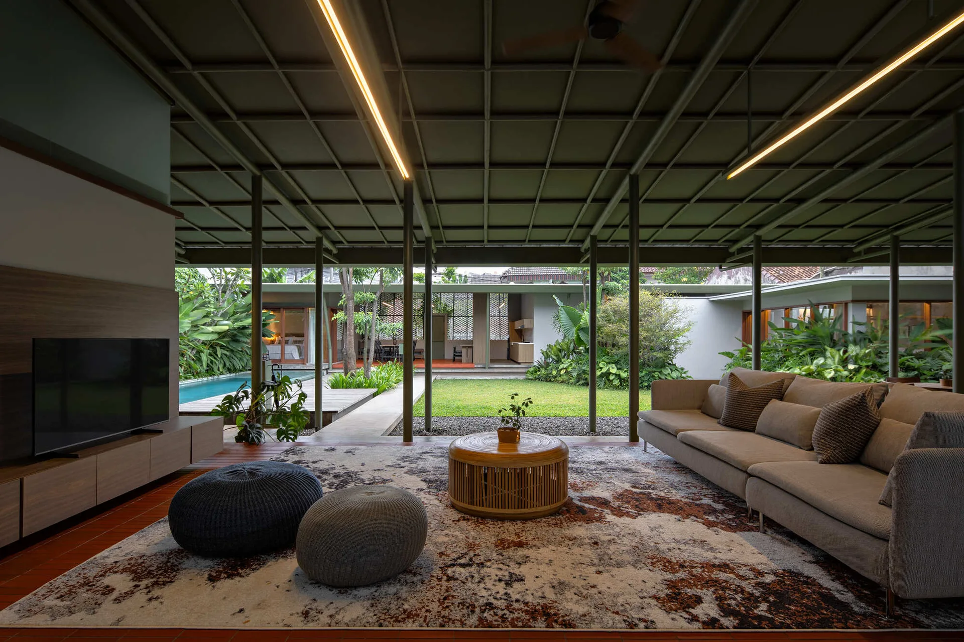 An open-air living room features a grey sectional sofa, an abstract patterned rug, and a low media console facing a lush garden and swimming pool.