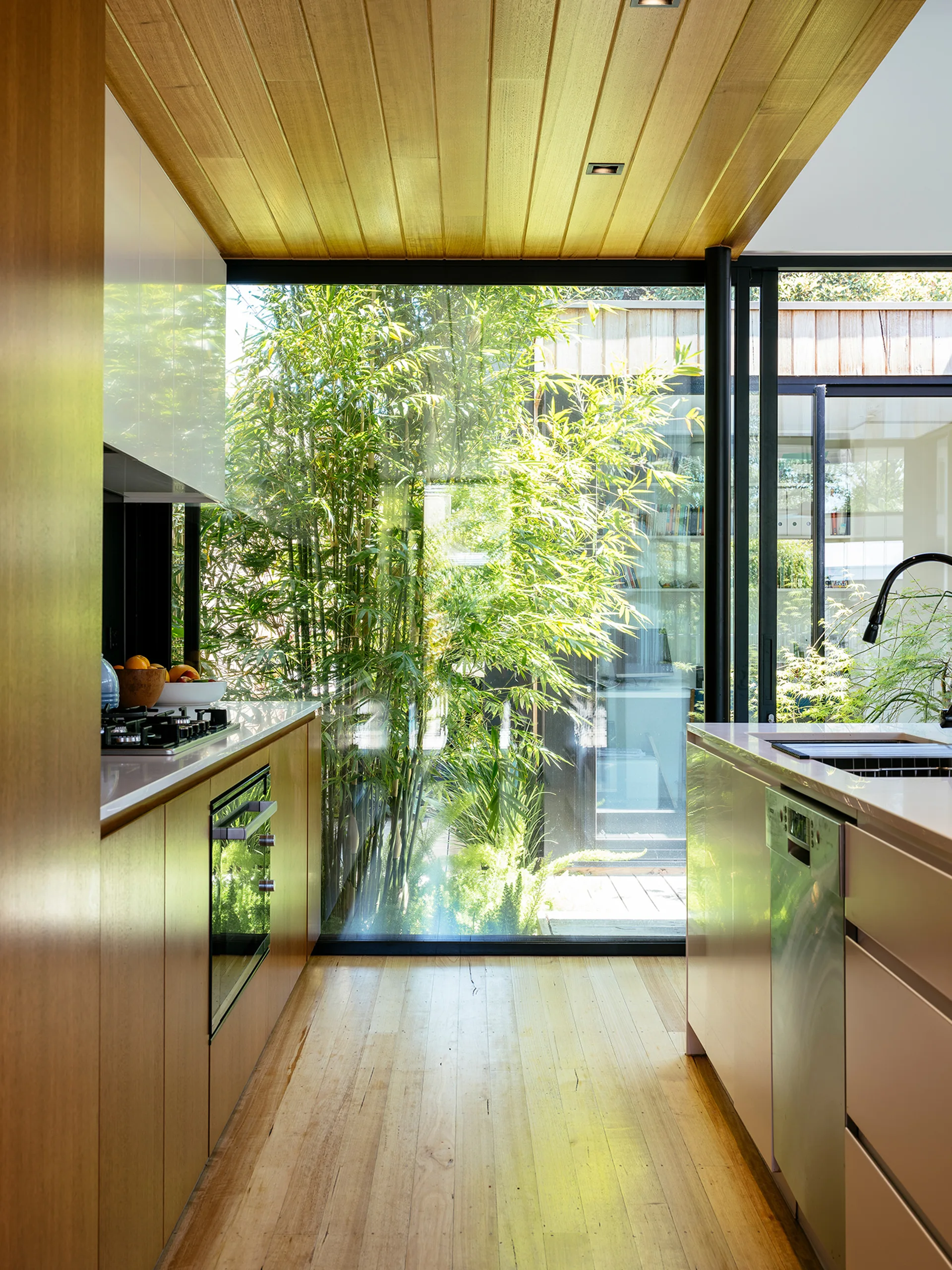 A modern galley kitchen features light wood cabinets, a wood plank ceiling, and a floor-to-ceiling glass window showcasing an outdoor bamboo garden.