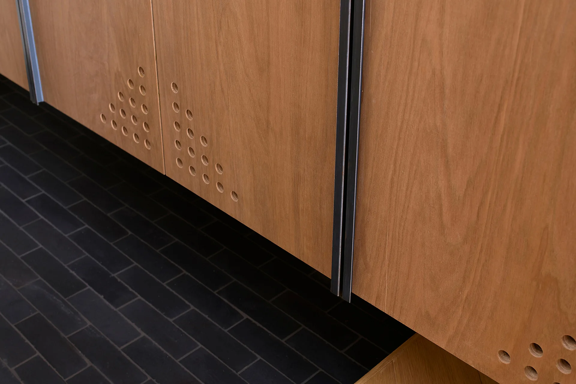 Close-up of wooden cabinet doors featuring a triangular pattern of small circular perforations and slim metal pulls over a dark tiled floor.