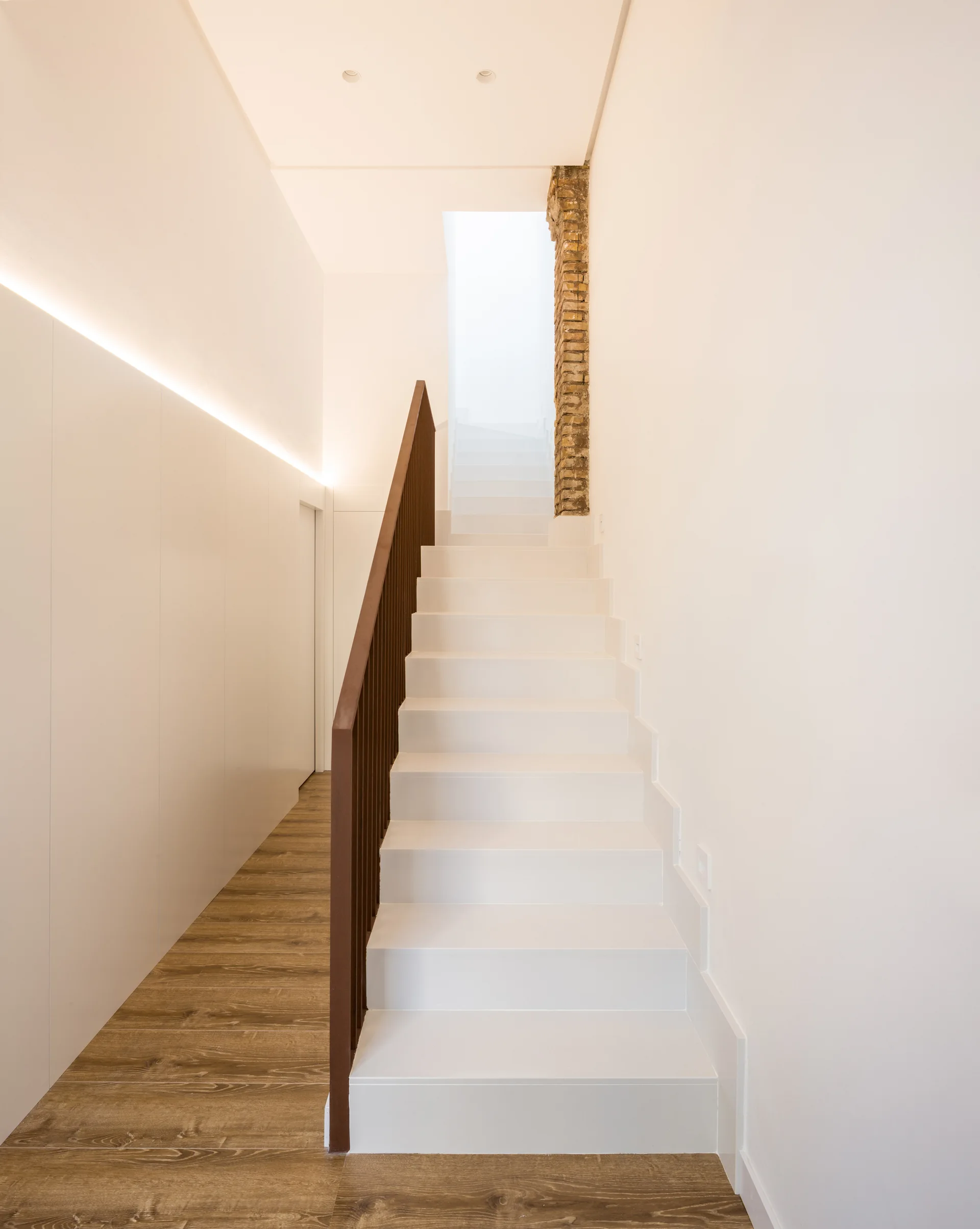 A minimalist white staircase features a dark metal handrail, wood flooring, a white paneled wall with integrated cove lighting, and an exposed brick accent.