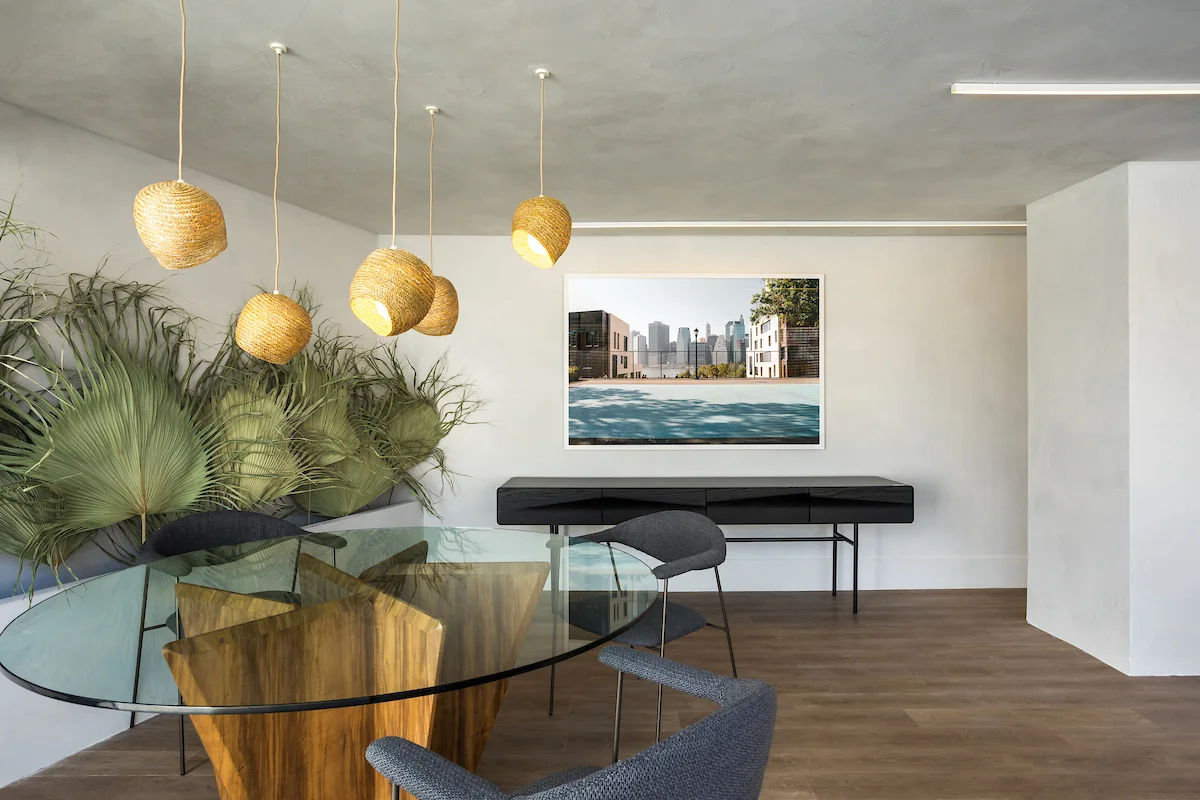 A dining room featuring a round glass table with a wooden base, dark grey chairs, woven pendant lights, and dried palm fronds against a plaster wall.