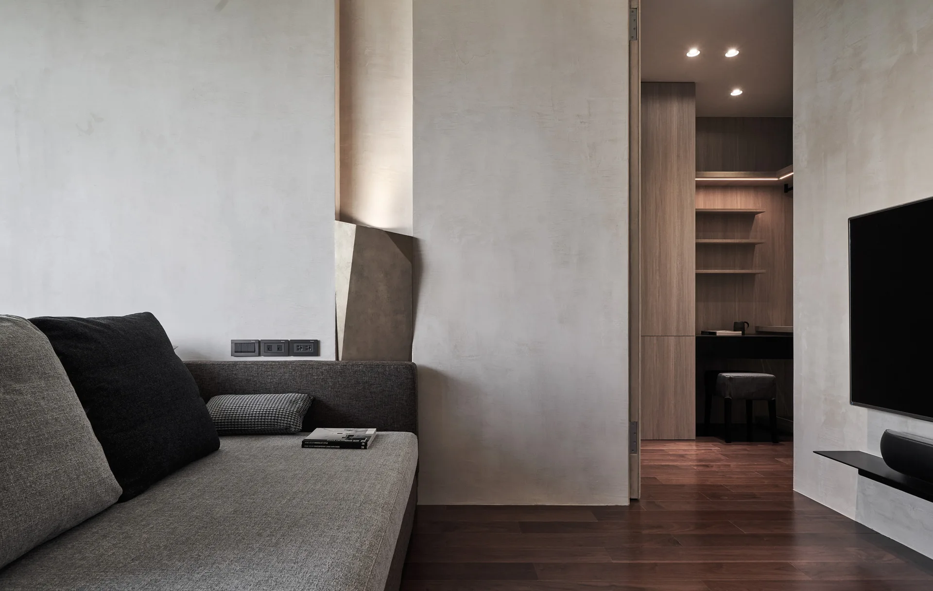 A minimalist living room features a grey sofa, textured plaster walls, and a wide doorway looking into a wood-paneled office nook.