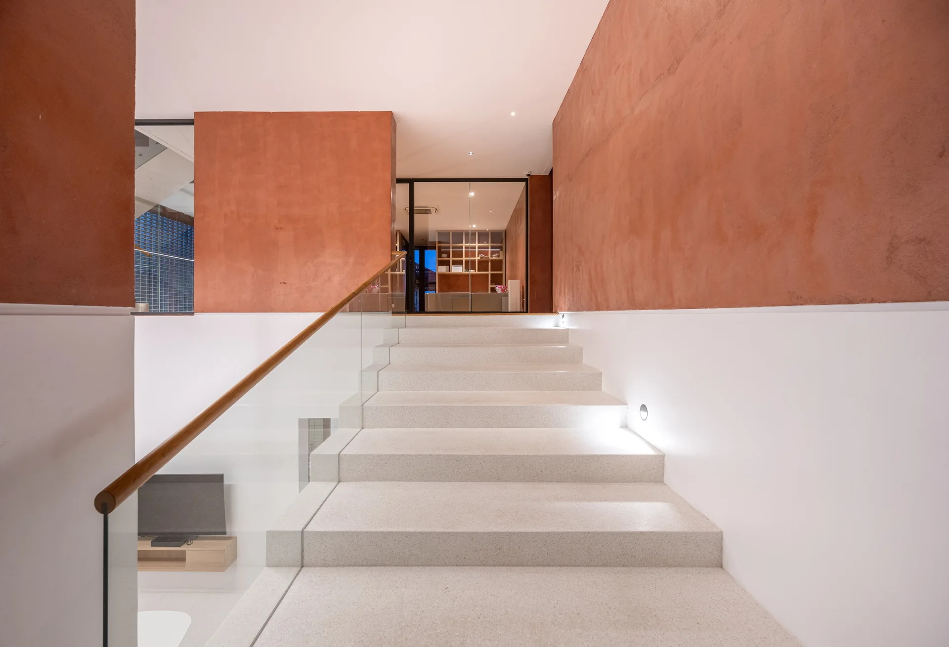 A modern stone staircase features a glass railing with a wood handrail, set against walls painted in terracotta and white.