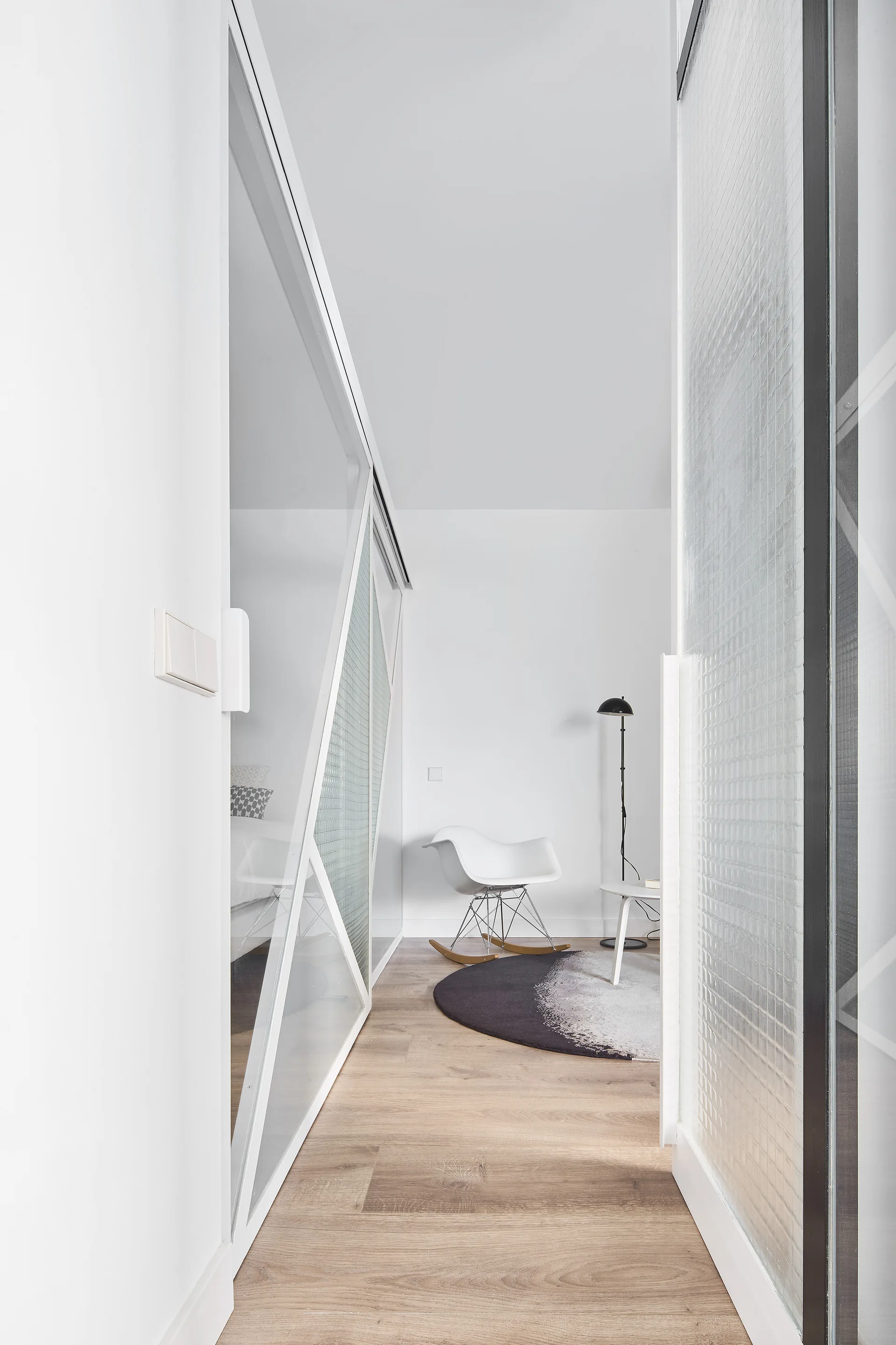 A bright hallway with wood floors features geometric glass sliding doors leading to a small nook with a white rocking chair and a round rug.