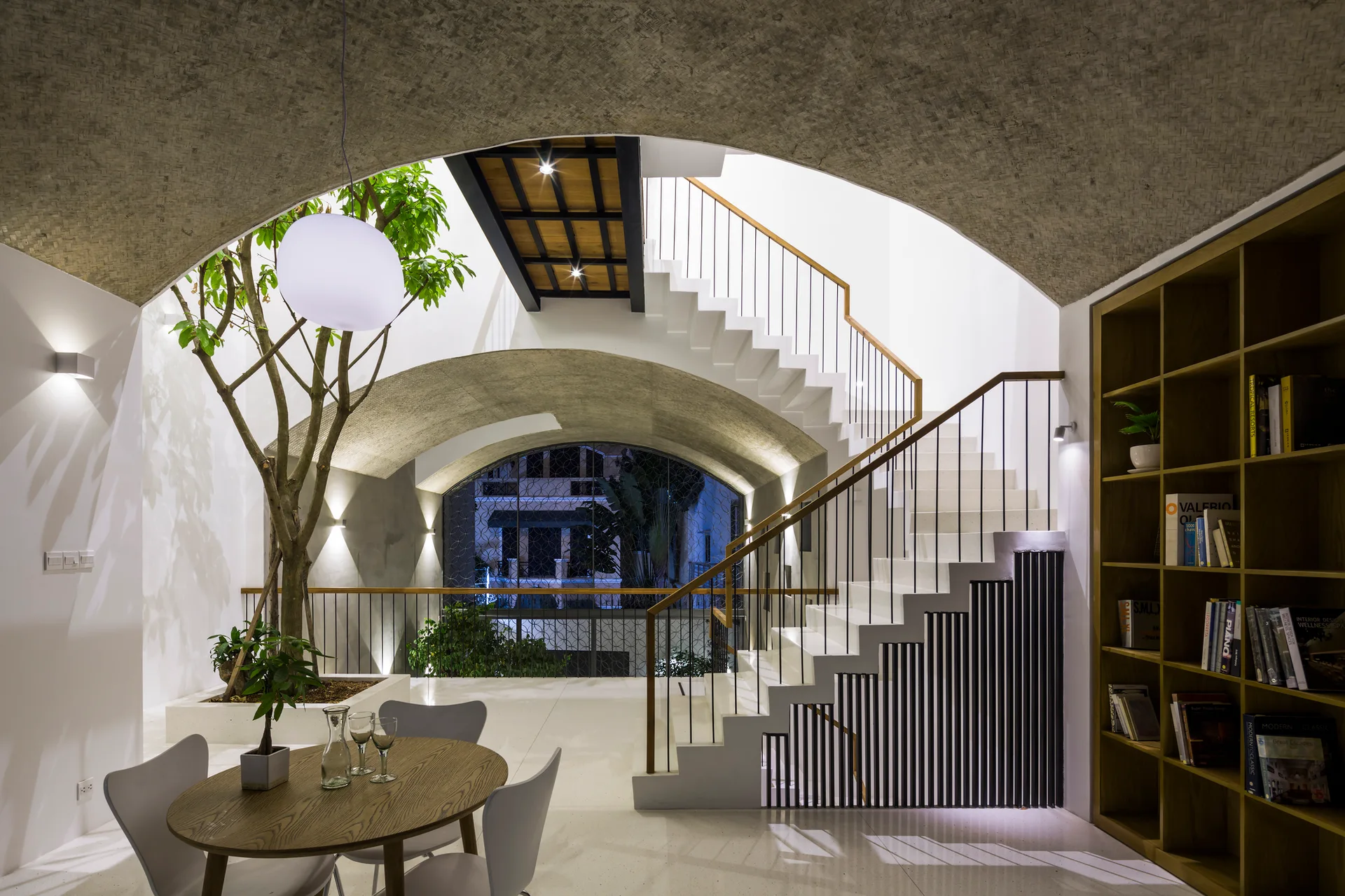A modern multi-level interior features a white concrete staircase, a textured barrel-vaulted ceiling, and a round wooden dining table.