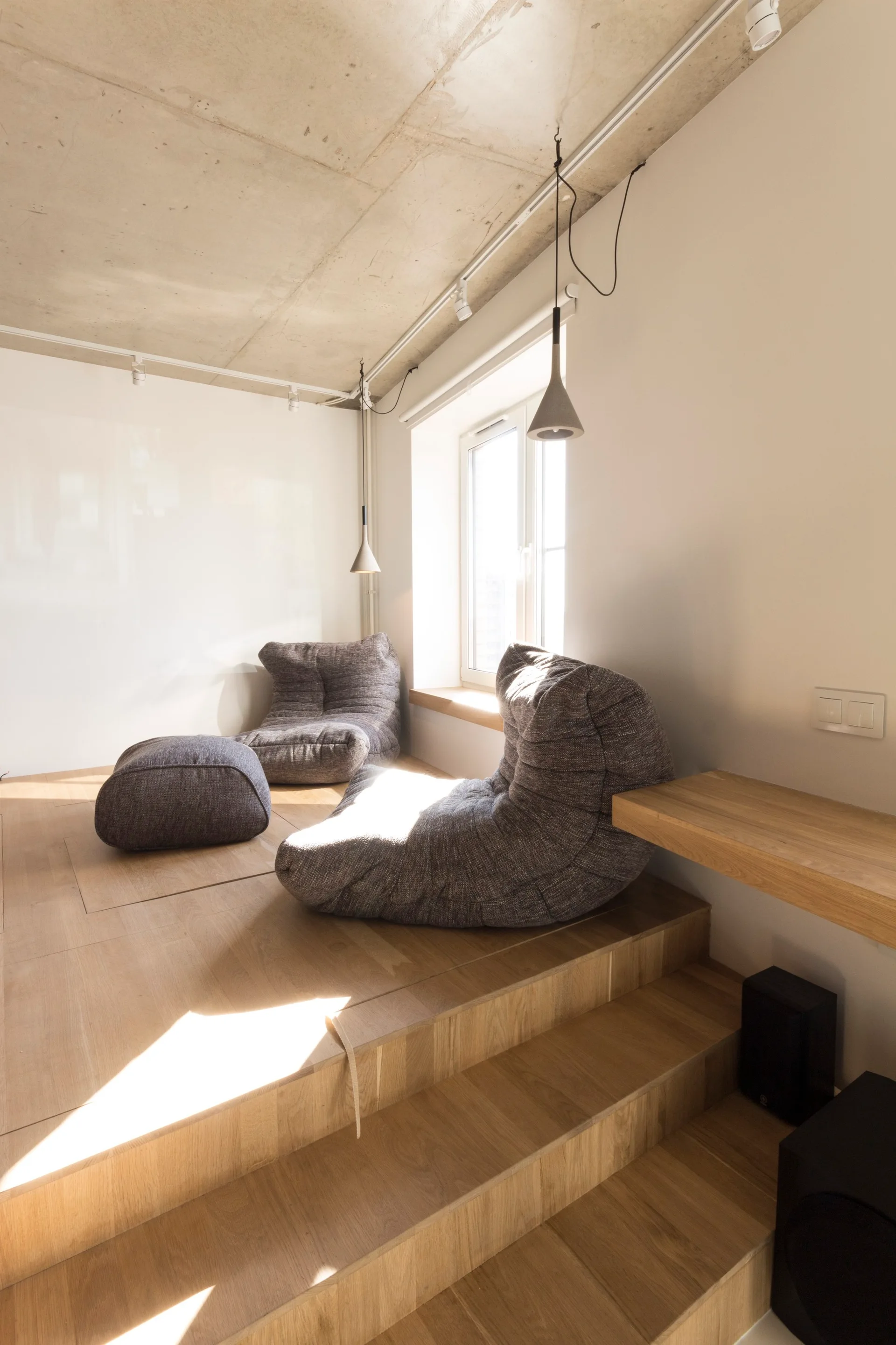 A modern lounge features grey textured slouchy chairs on a raised light wood platform under an exposed concrete ceiling with hanging pendant lights.