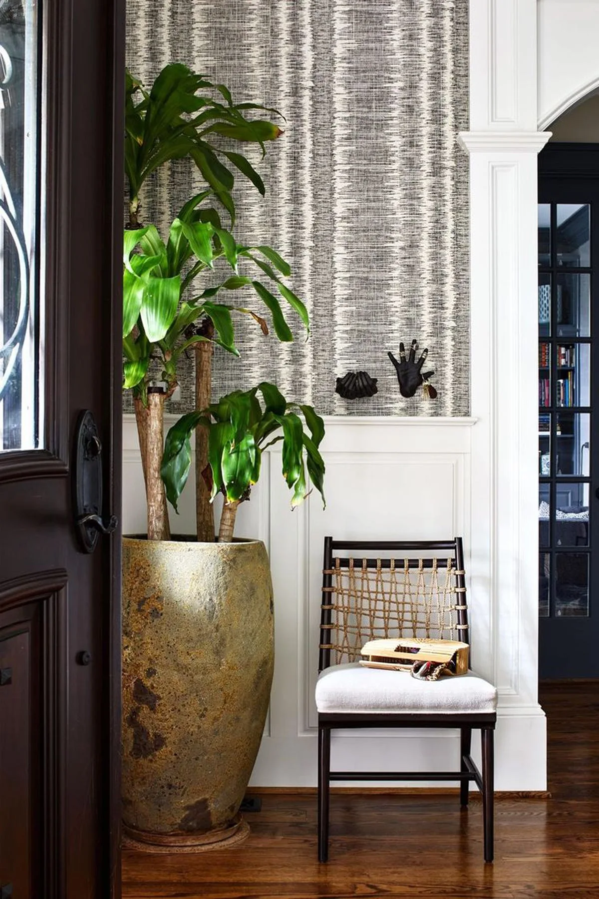 An entryway featuring white wainscoting, textured grey and white wallpaper, a tall plant in a large stone planter, and a woven back chair.