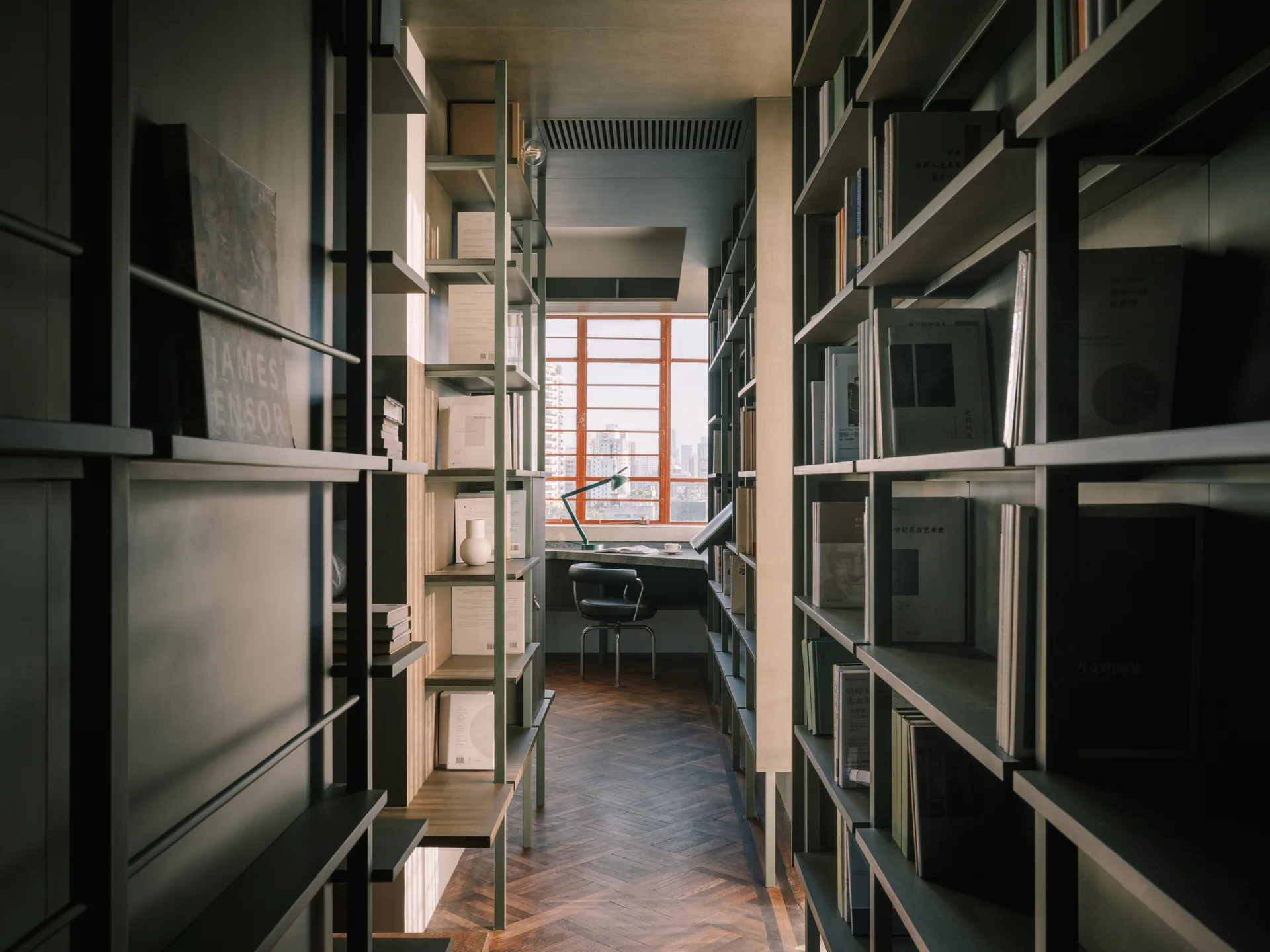 A narrow corridor lined with tall dark green metal bookshelves on a herringbone wood floor leading to a window with a built-in desk and chair.