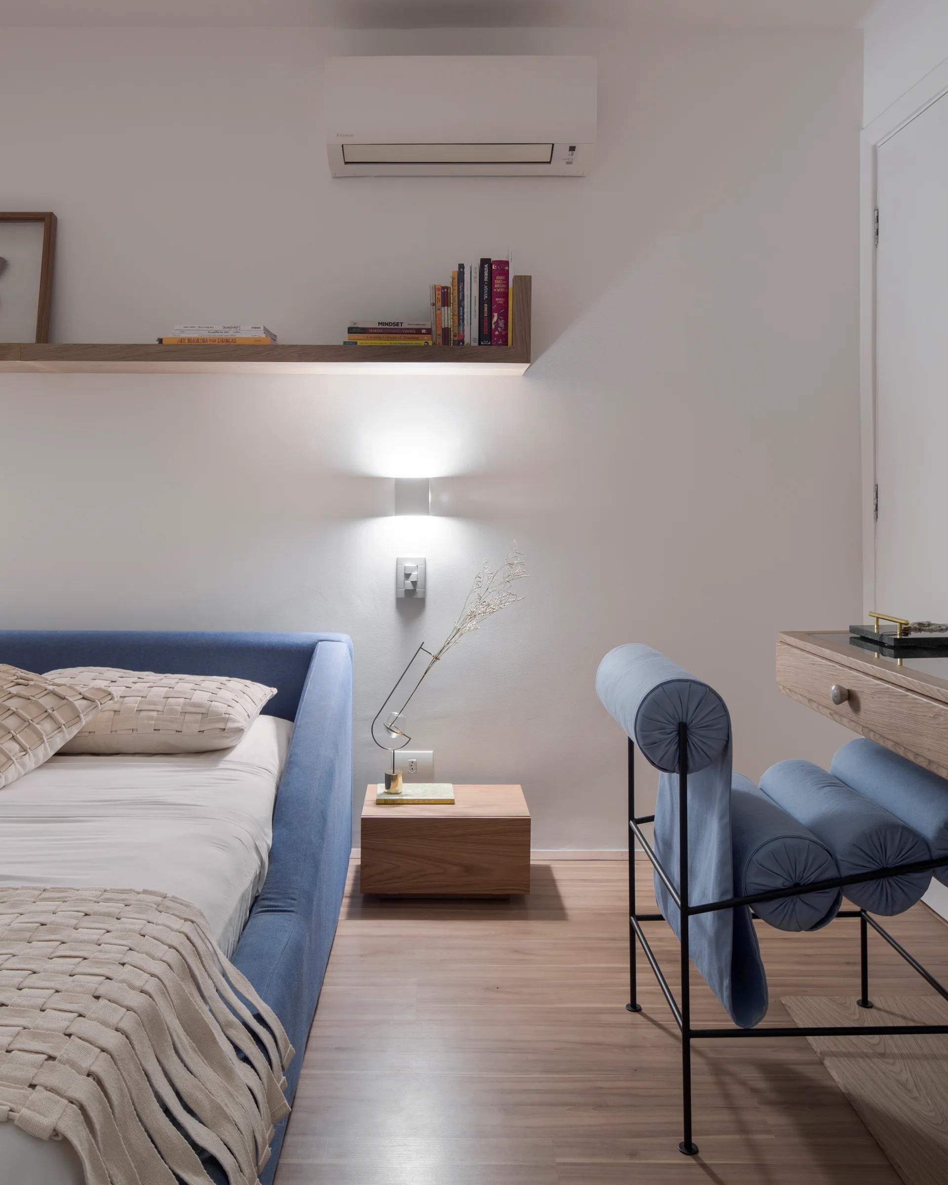 A modern bedroom features a blue upholstered bed, a matching blue bolster chair, a low wooden nightstand, and a floating shelf on a white wall.