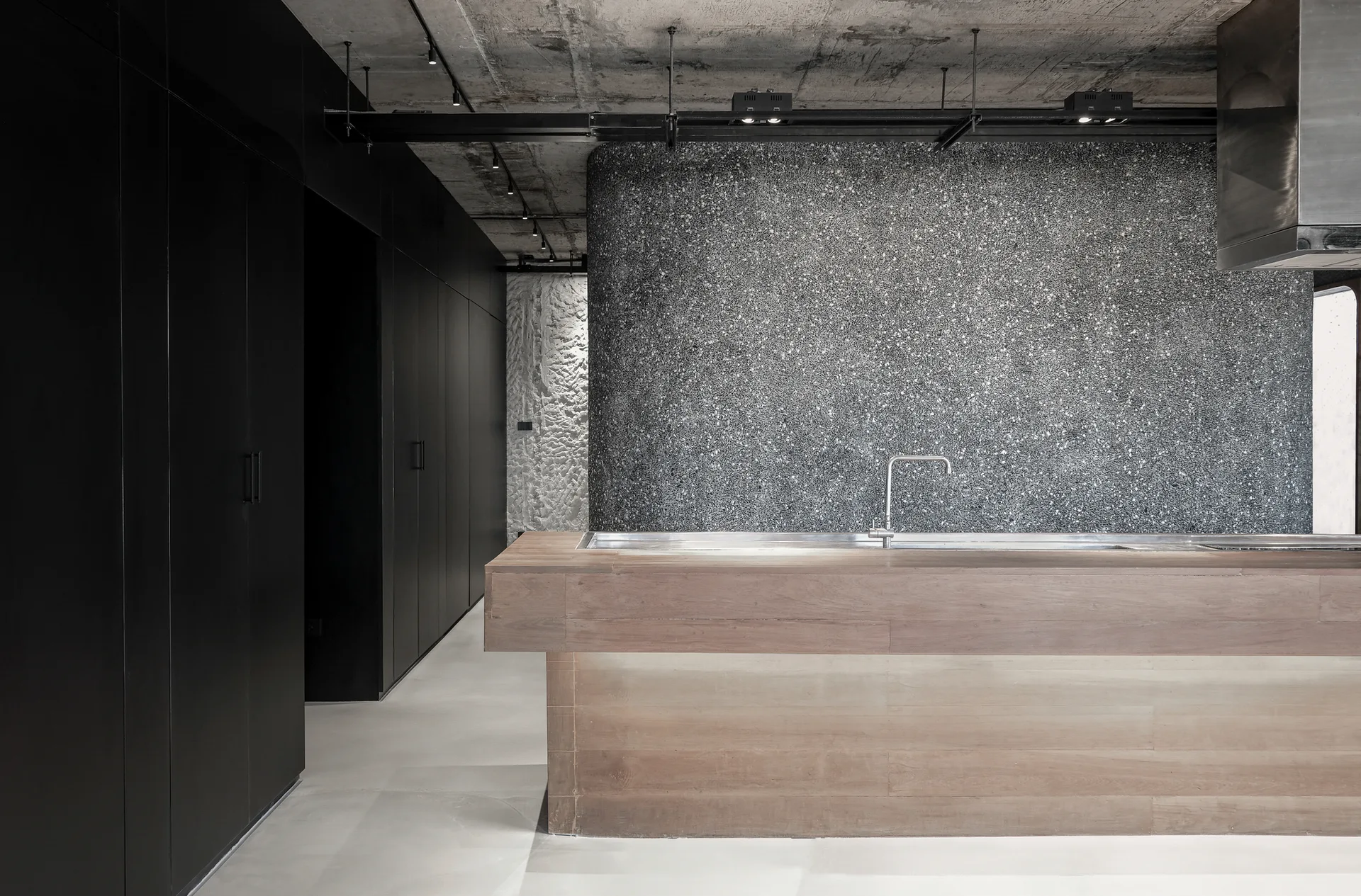 A modern industrial kitchen featuring a wooden island with a silver faucet set against a curved speckled grey wall and matte black cabinets.