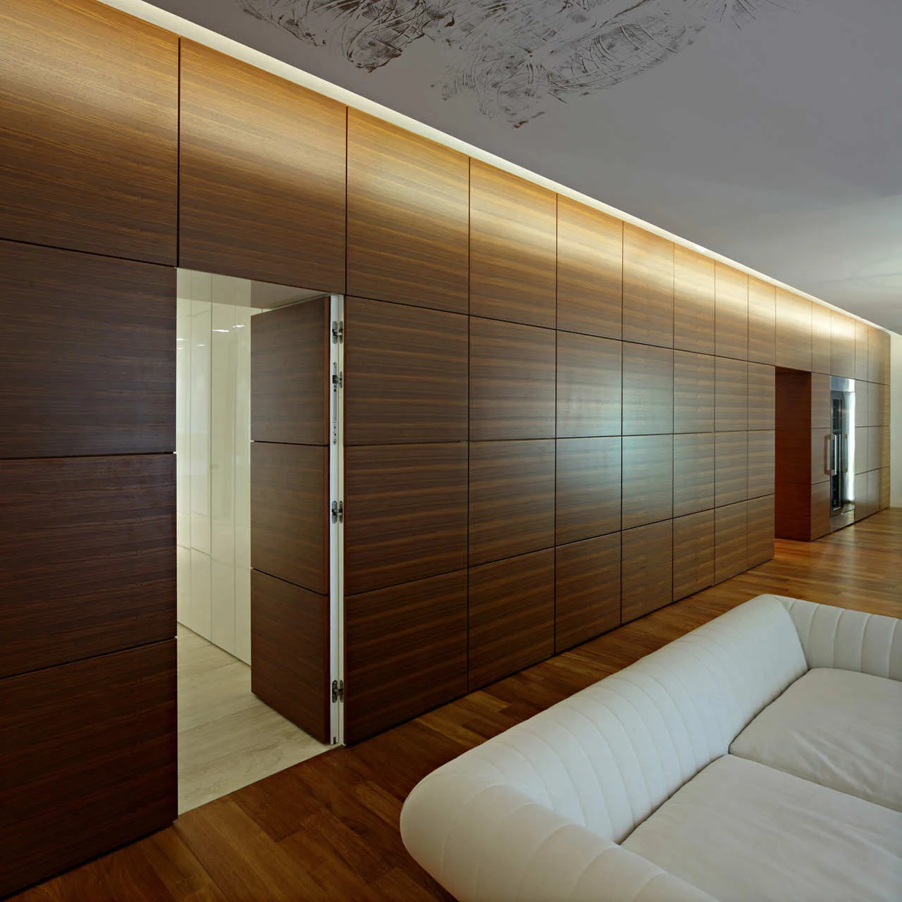 A white sofa sits opposite a continuous dark wood paneled wall with an open integrated door and indirect cove lighting along the ceiling.