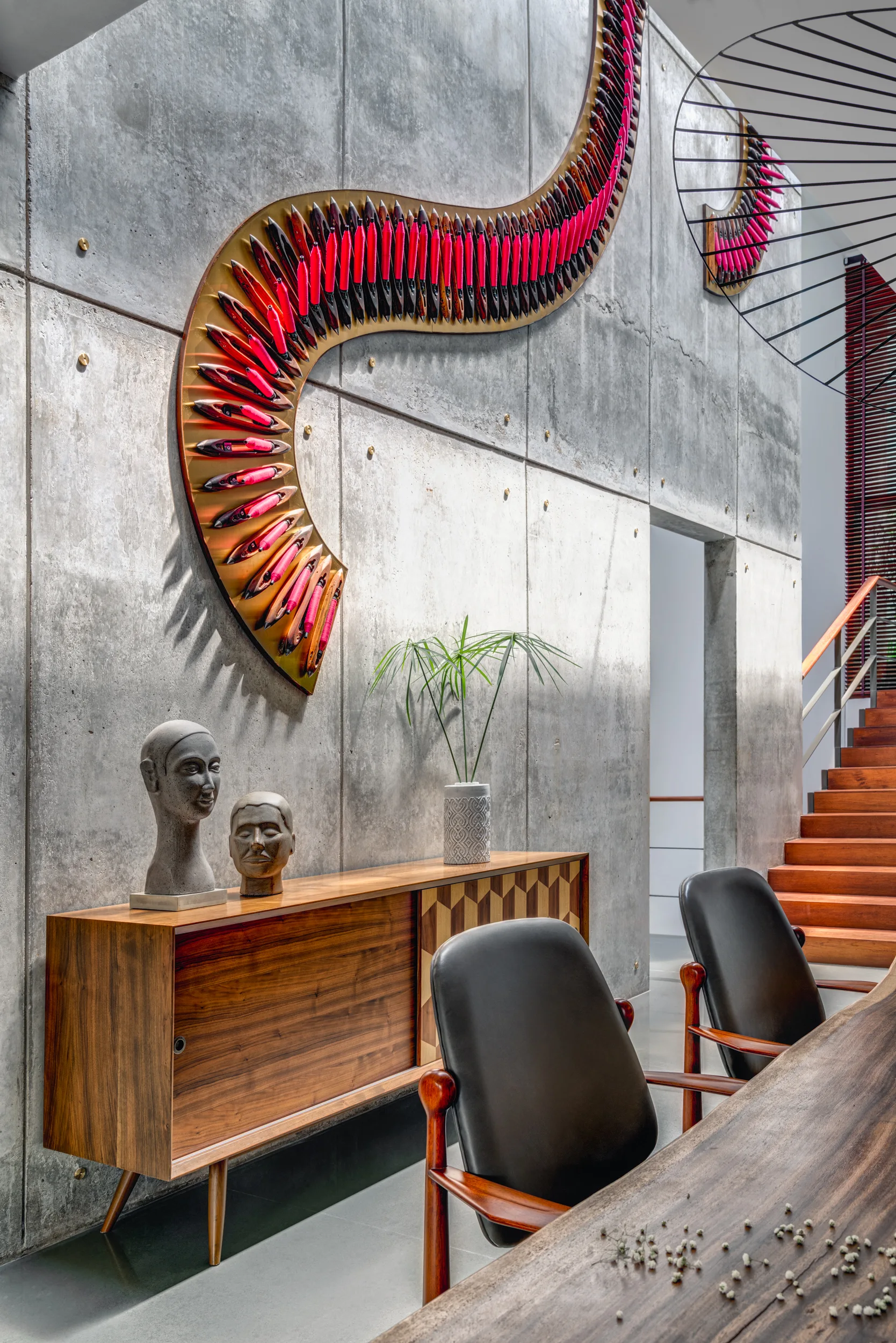 A modern dining area featuring a raw concrete wall adorned with a pink curving sculpture, a walnut wood credenza, and black leather chairs.