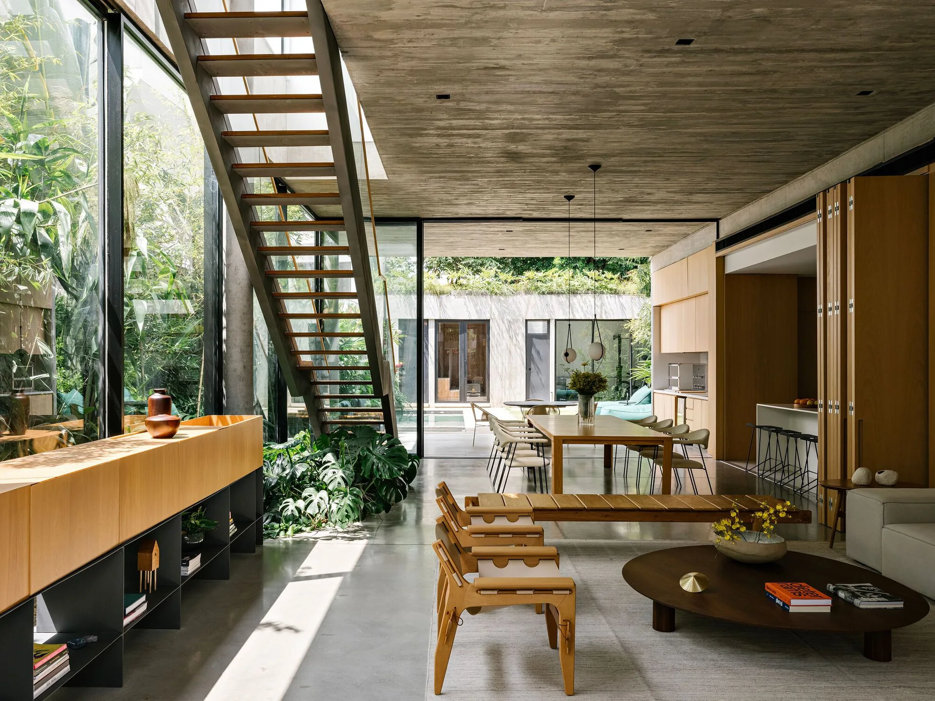A modern open-plan living room featuring board-formed concrete ceilings, an open-tread metal staircase, large glass walls, and low wood furniture.