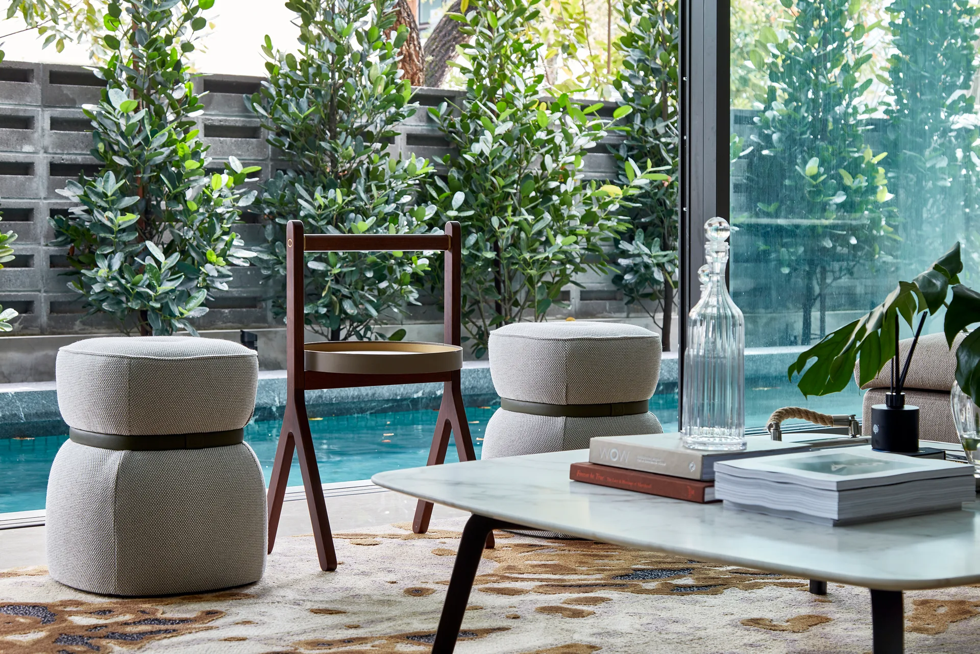 A modern living room features a marble coffee table, two textured grey ottomans, and a wooden chair in front of a glass wall overlooking a swimming pool.