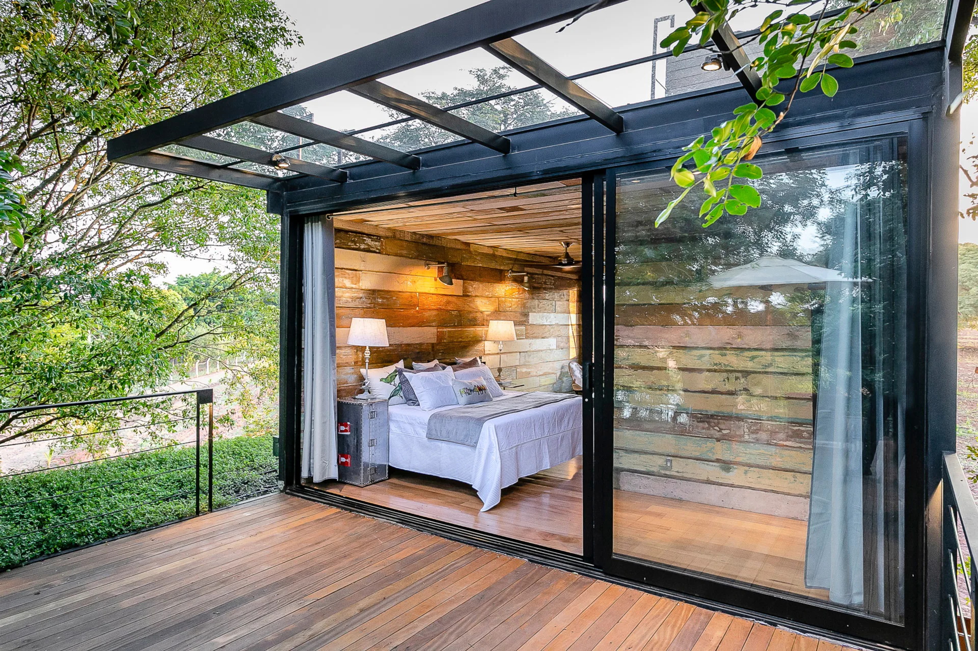 A rustic bedroom with wood-plank walls and a bed is viewed through large sliding glass doors leading to an outdoor wooden deck.