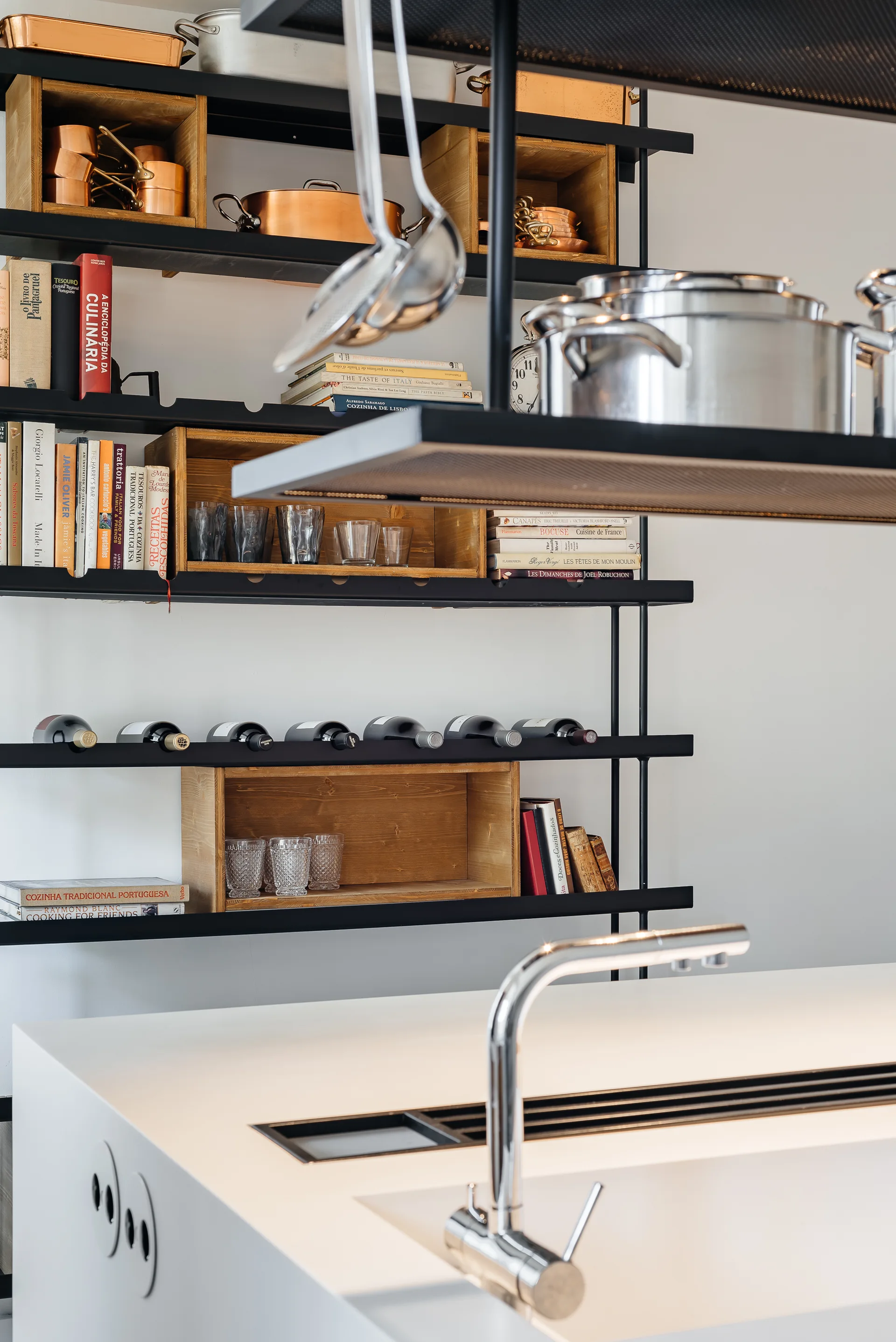 A modern kitchen details black metal open shelving with wooden box inserts holding cookbooks, copper pots, and glassware above a white island.