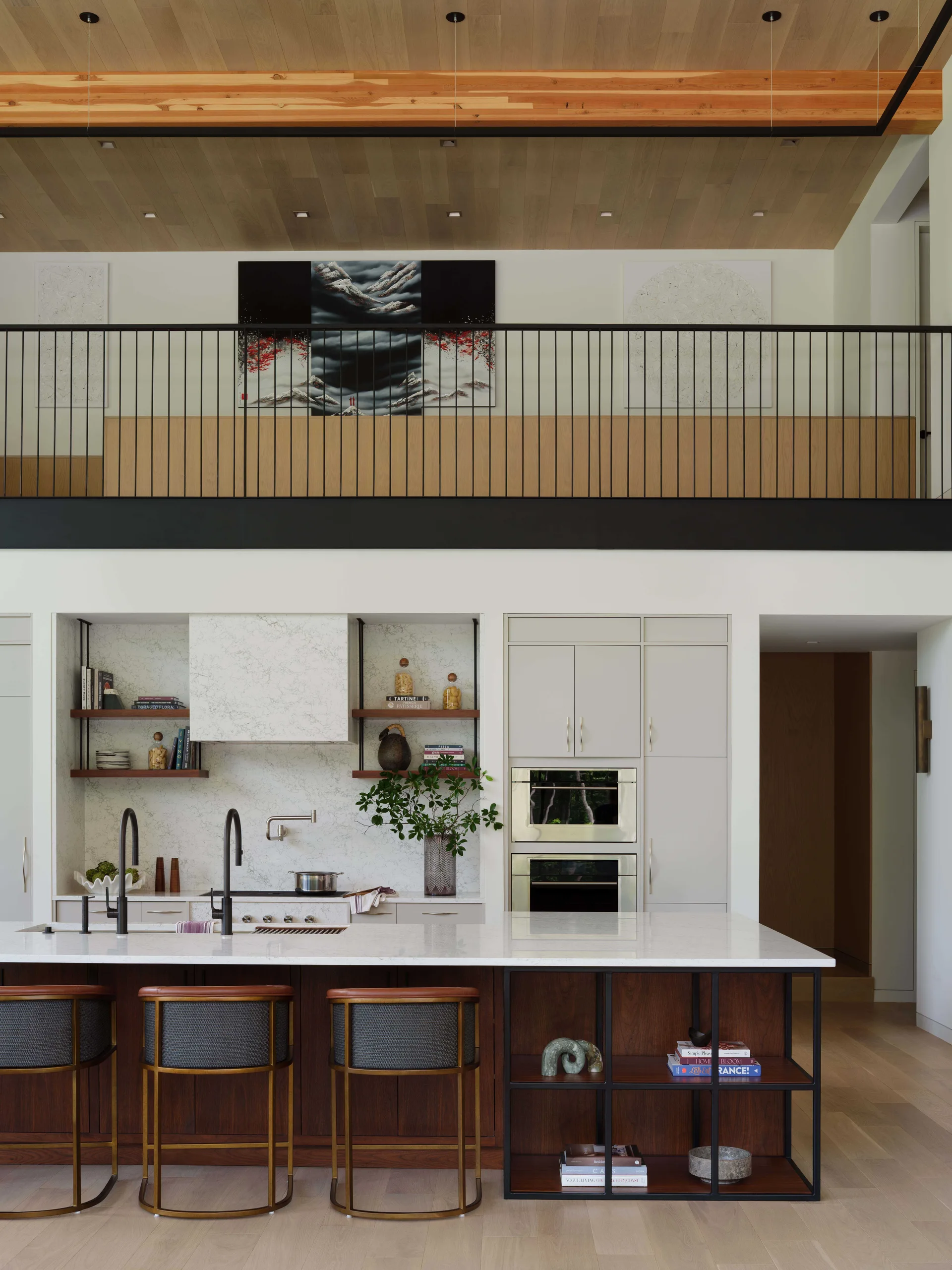 A modern kitchen features a marble-topped island with brass stools beneath a wood-paneled ceiling and an upper mezzanine balcony with black metal railing.