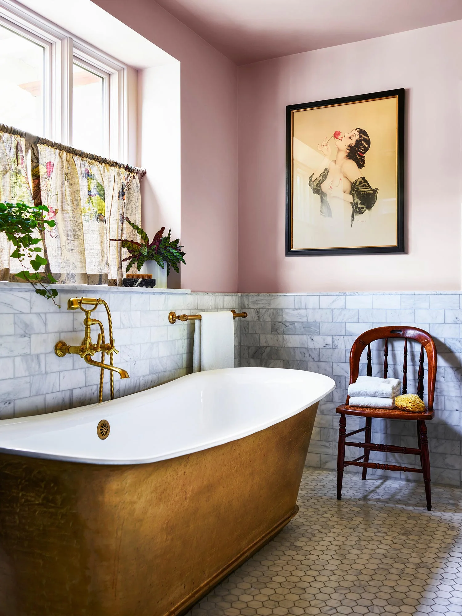 A gold-toned freestanding bathtub sits below a window in a bathroom with pink walls and white marble subway tile.
