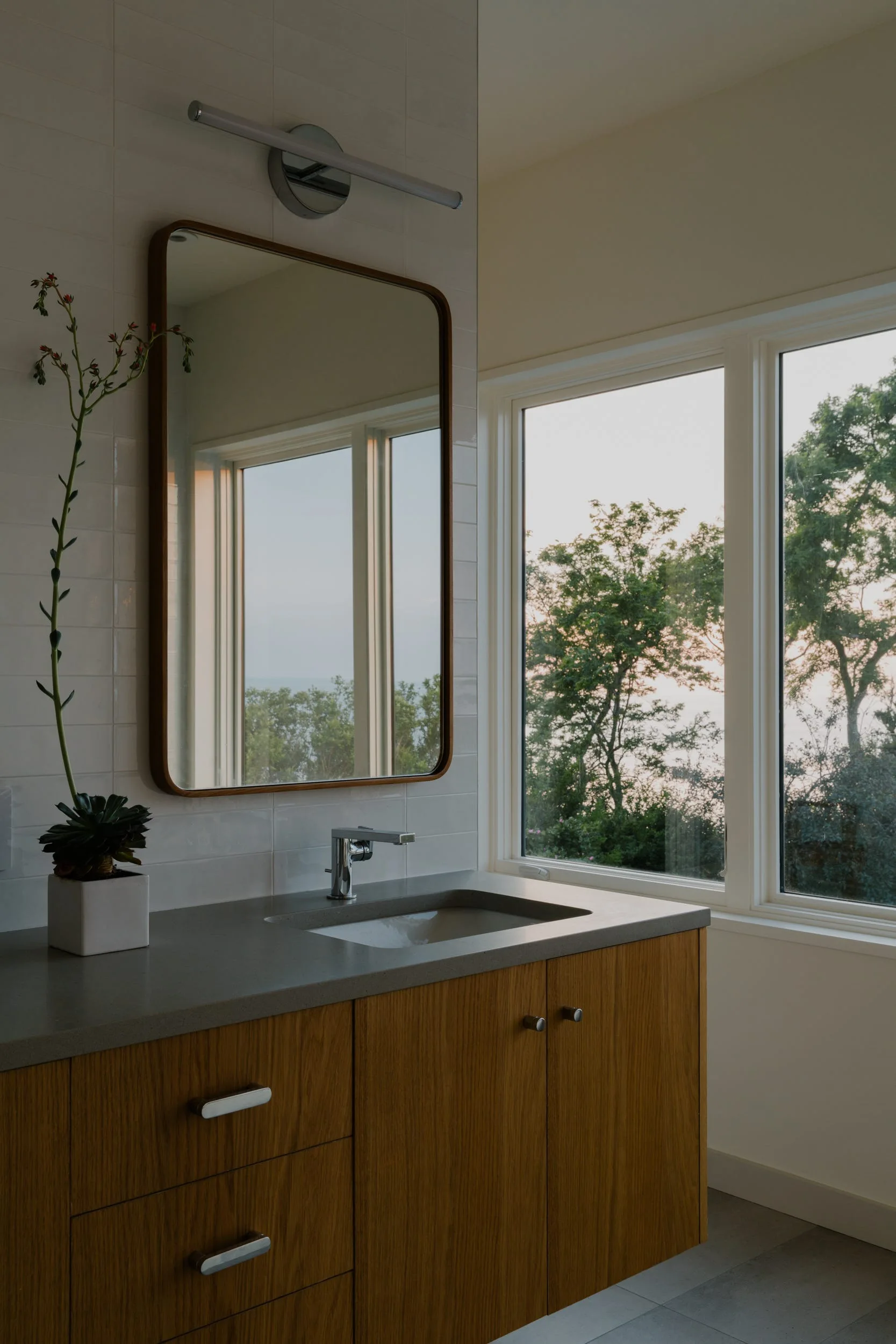 A modern bathroom features a wood floating vanity with a grey countertop, a chrome faucet, a rounded rectangular mirror, and a window with outdoor views.