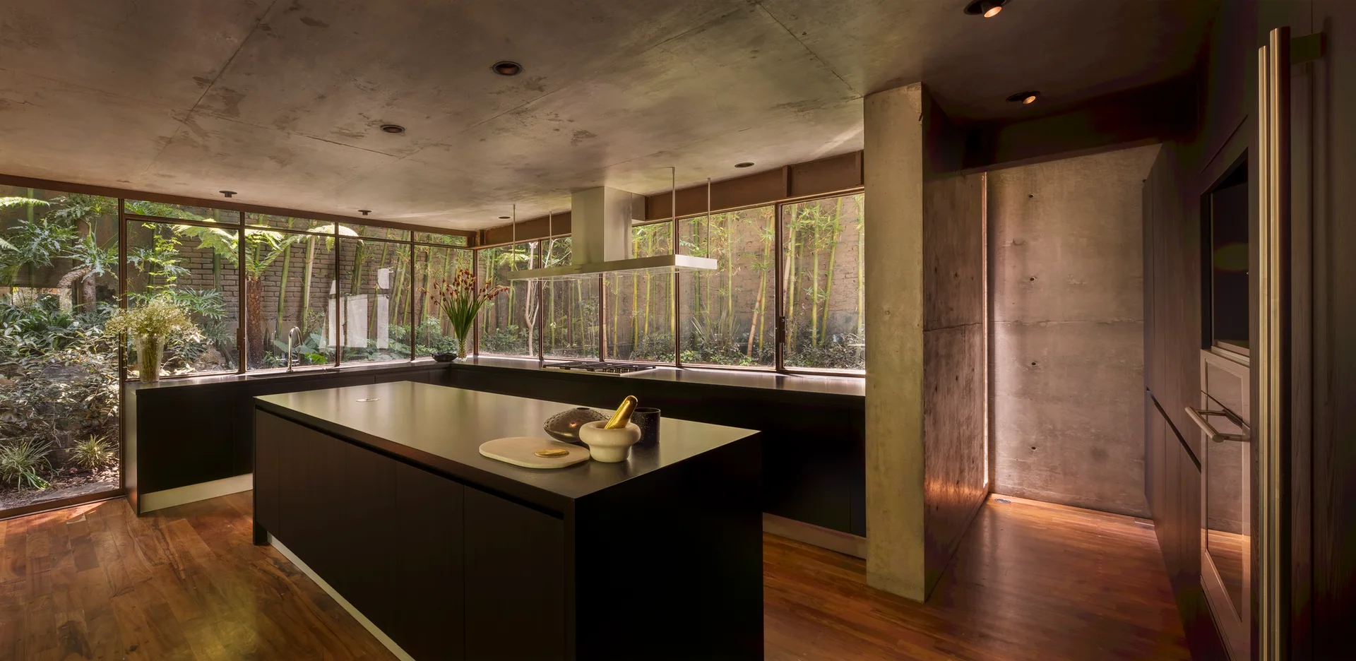 A modern kitchen features a black island, matte black cabinets, wood floors, a concrete ceiling, and large windows looking out to a garden.