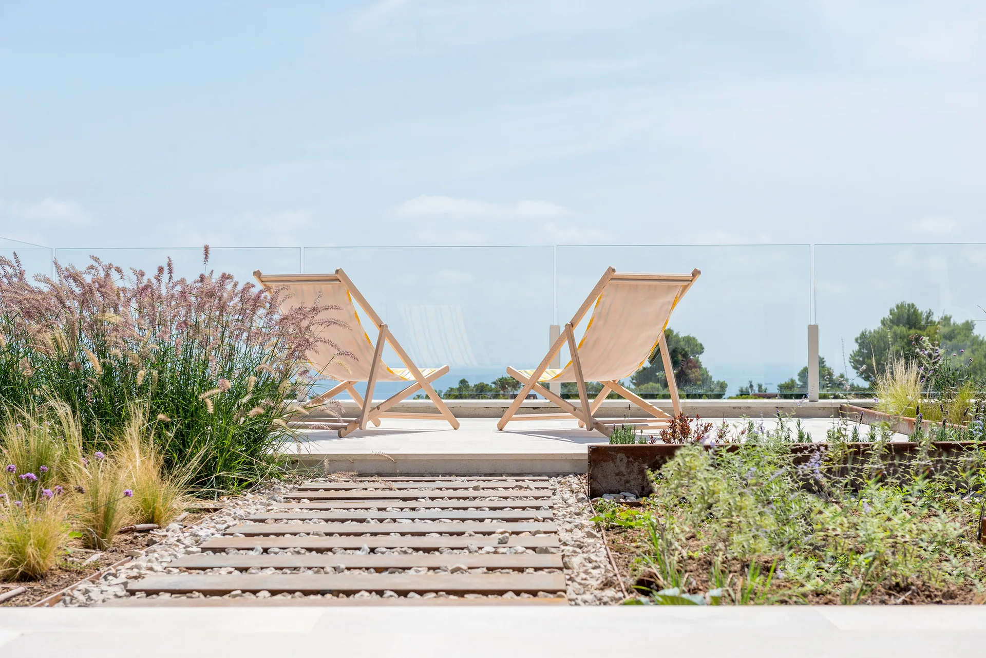 Two wooden folding lounge chairs sit on a stone terrace behind a glass railing, with a gravel and timber pathway leading through wild grasses.