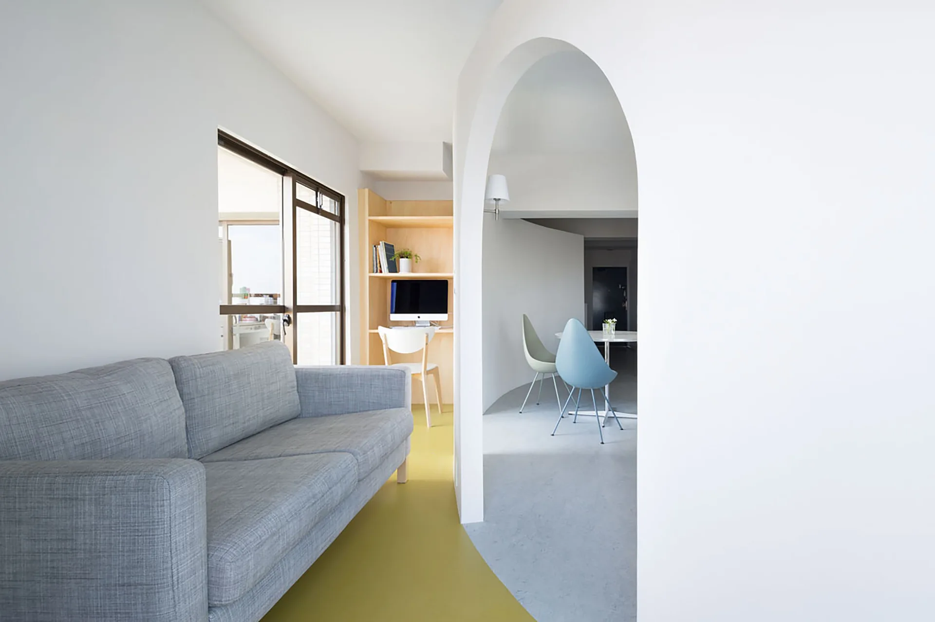 A minimalist living space featuring a grey sofa, yellow flooring, and a white arched doorway that leads to a dining area.