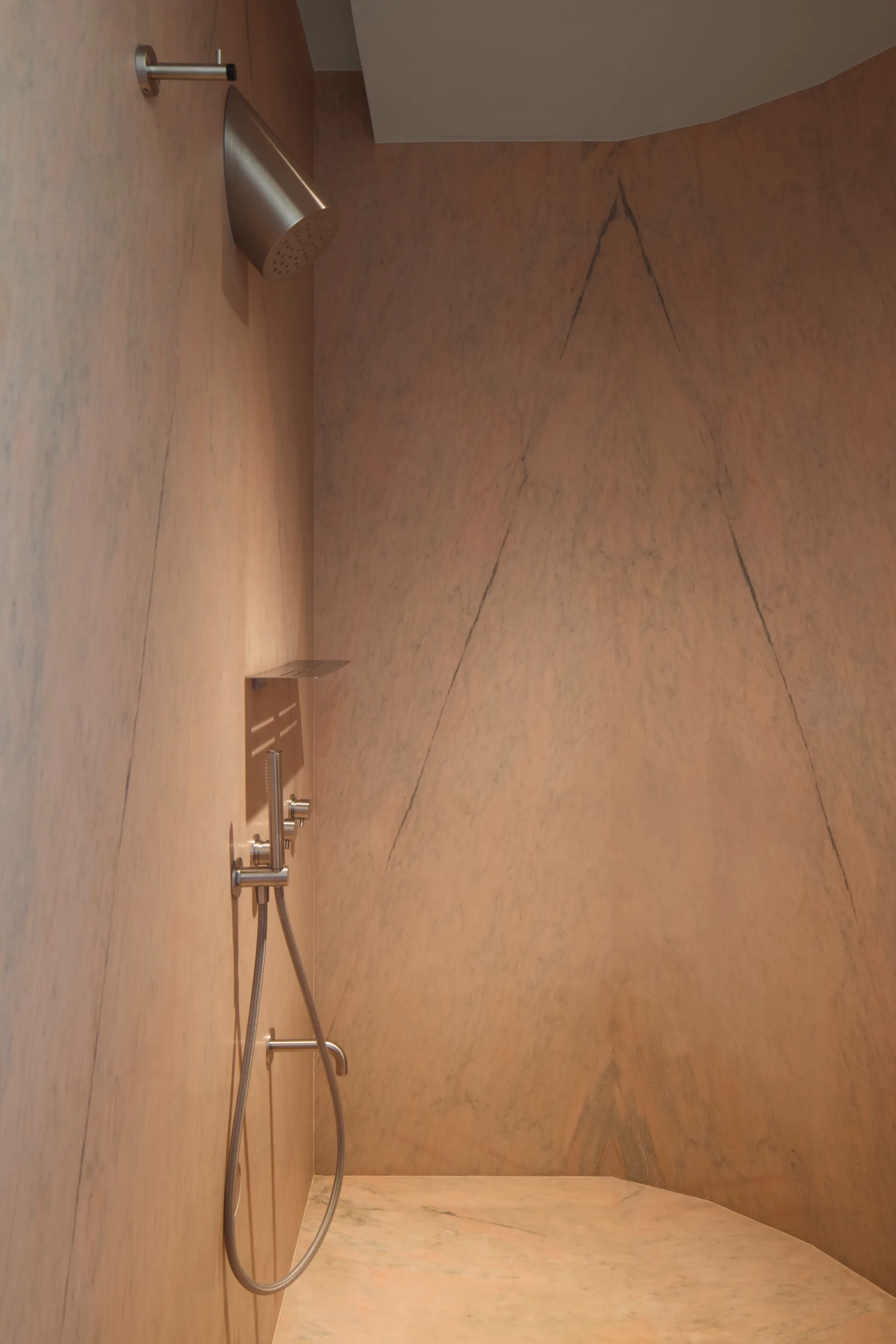 A modern walk-in shower with veined pink marble walls, brushed silver hardware, a handheld sprayer, and a small metal shelf.
