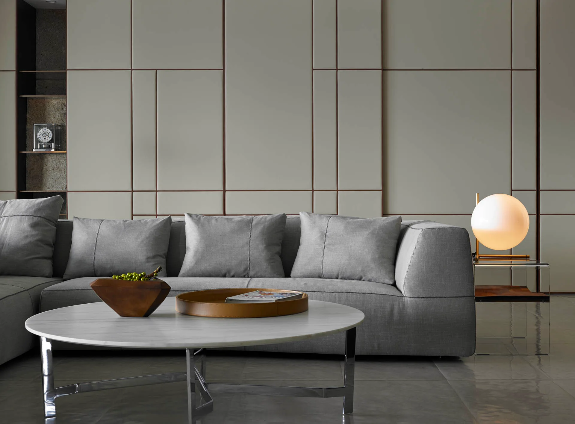 A grey upholstered sofa sits before a geometric paneled wall next to a round marble coffee table and a glowing globe table lamp.